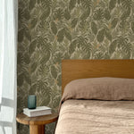 Bed with tan linens and wooden headboard, wooden side table  with white book and green glass, white floor length curtain against a olive green and tan tropical wallpaper. 