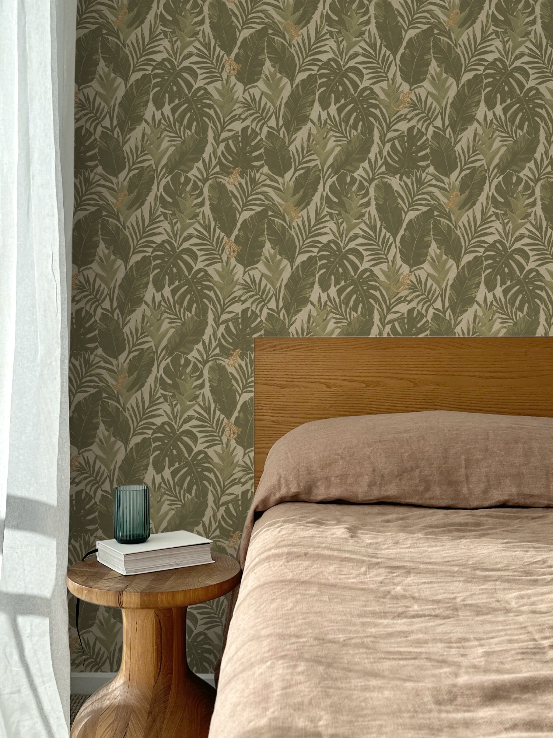 Bed with tan linens and wooden headboard, wooden side table  with white book and green glass, white floor length curtain against a olive green and tan tropical wallpaper. 