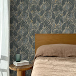 Bed with tan linens and wooden headboard, wooden side table  with white book and green glass, white floor length curtain against a steel blue and tan tropical wallpaper. 