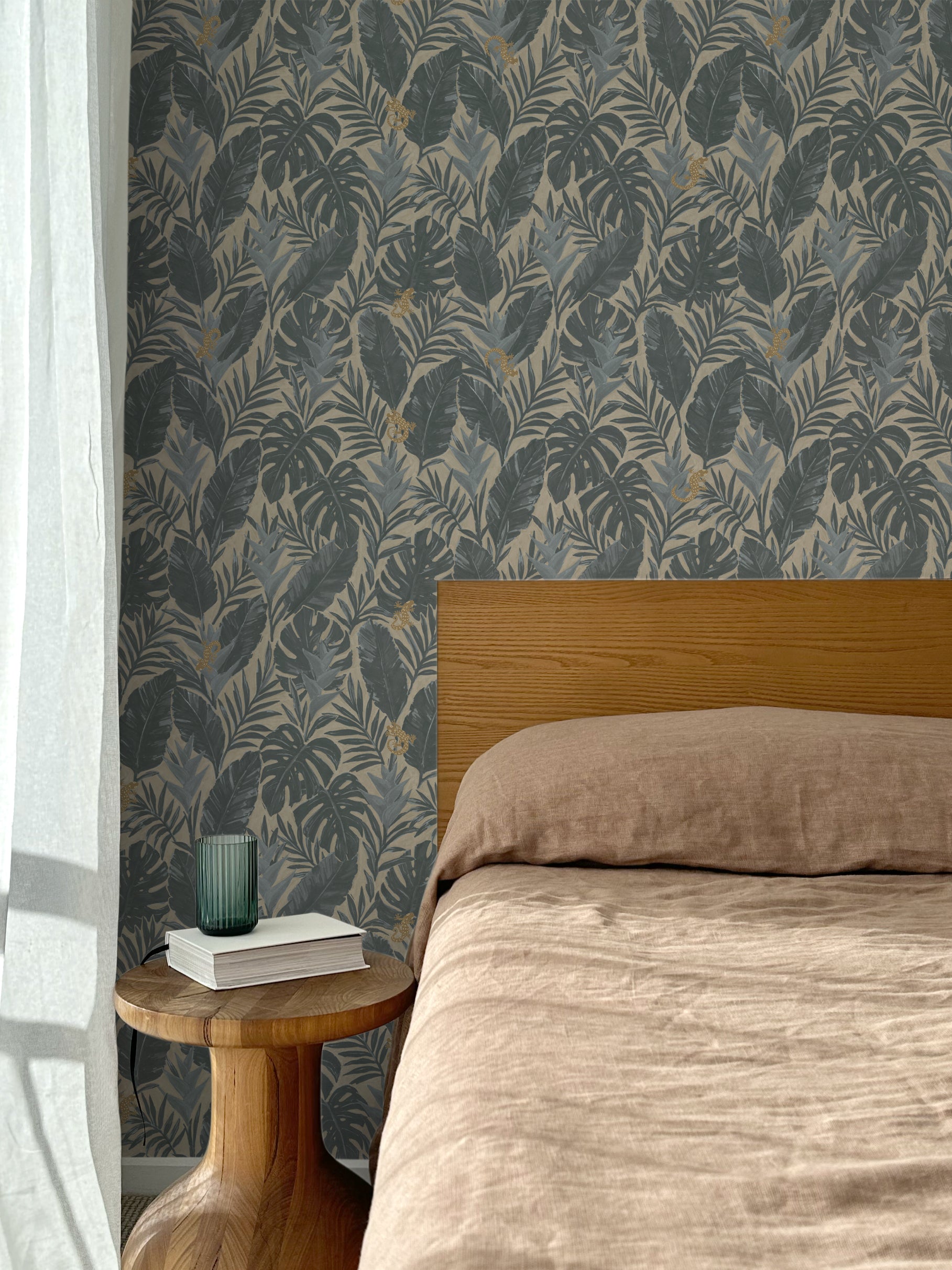 Bed with tan linens and wooden headboard, wooden side table  with white book and green glass, white floor length curtain against a steel blue and tan tropical wallpaper. 