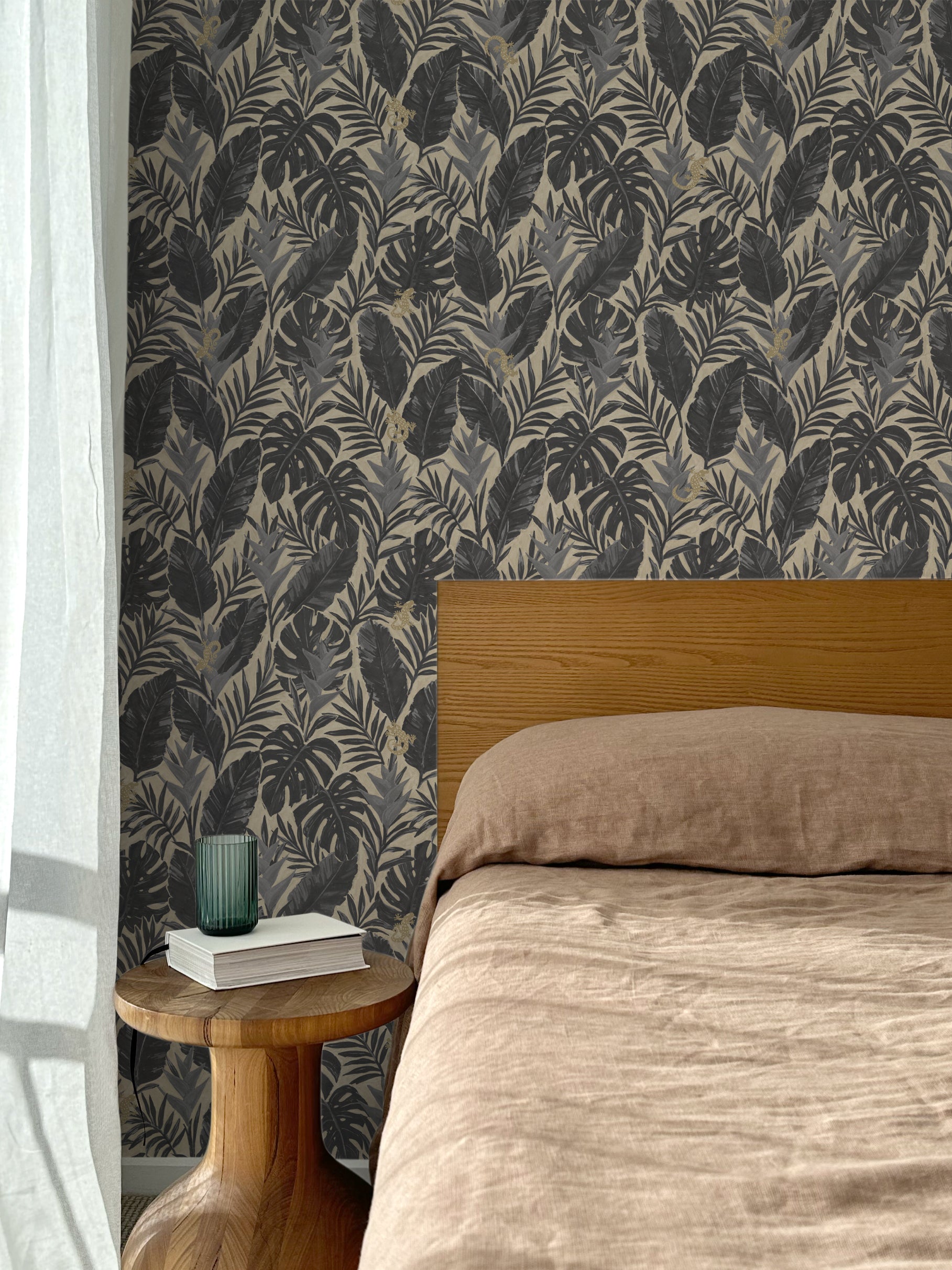 Bed with tan linens and wooden headboard, wooden side table  with white book and green glass, white floor length curtain against a charcoal grey and tan tropical wallpaper. 
