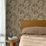 Bed with tan linens and wooden headboard, wooden side table  with white book and green glass, white floor length curtain against a taupe tropical palm frond wallpaper. 