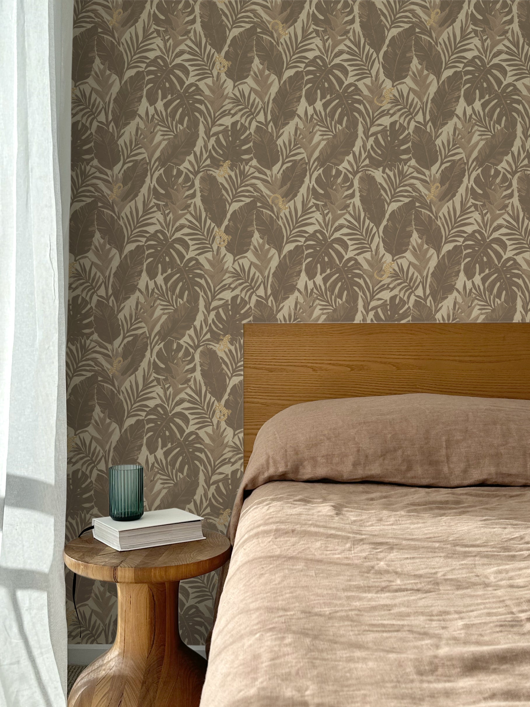Bed with tan linens and wooden headboard, wooden side table  with white book and green glass, white floor length curtain against a taupe tropical palm frond wallpaper. 