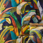 Wooden rattan chair with a bold painterly palm frond throw pillow in blue, yellow and red against matching wallpaper. 