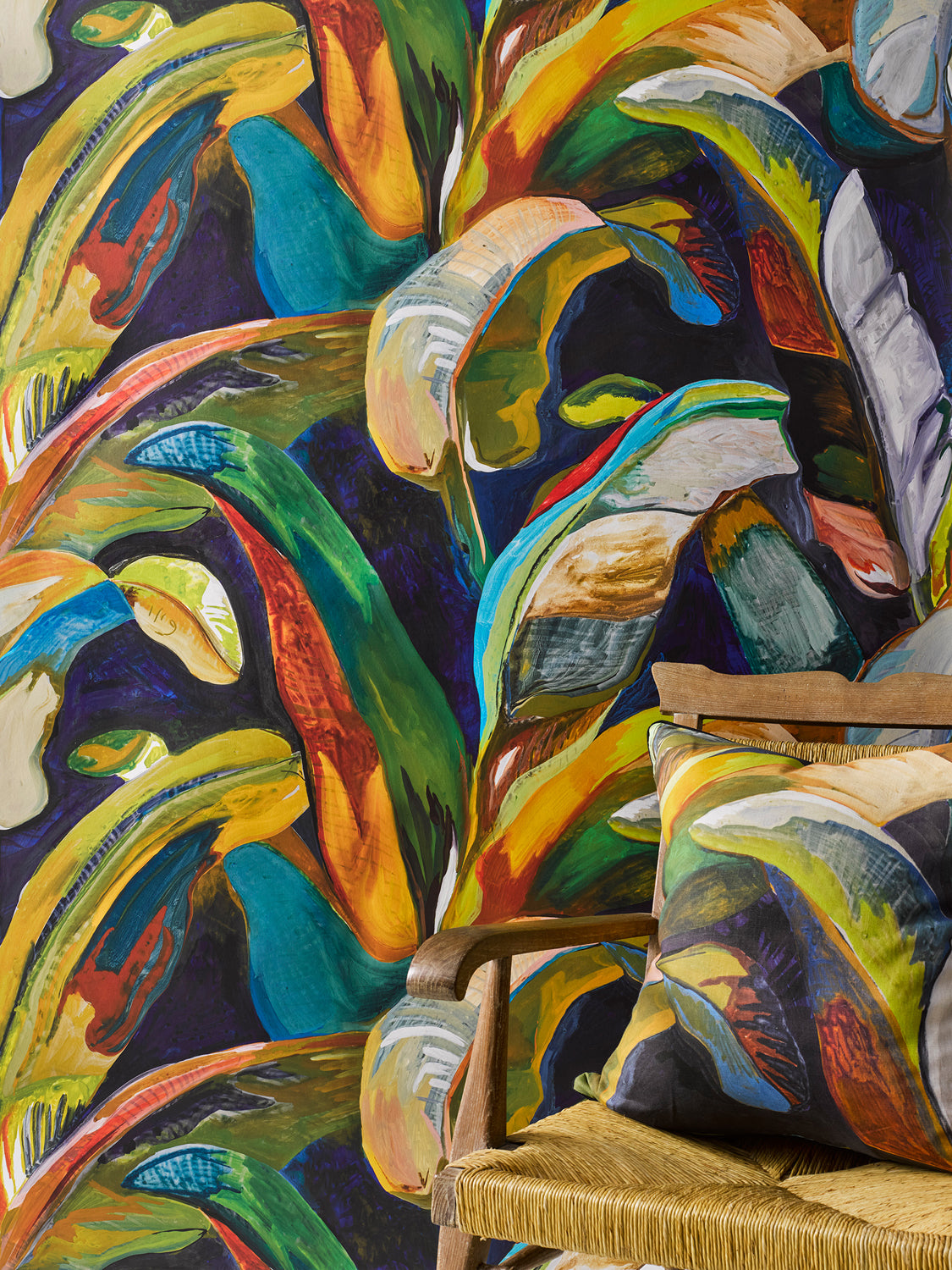 Wooden rattan chair with a bold painterly palm frond throw pillow in blue, yellow and red against matching wallpaper. 