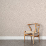 Angled wooden chair with woven seat on concrete floor against wallpaper in a textural wicker pattern in muted blush pink on a white field.