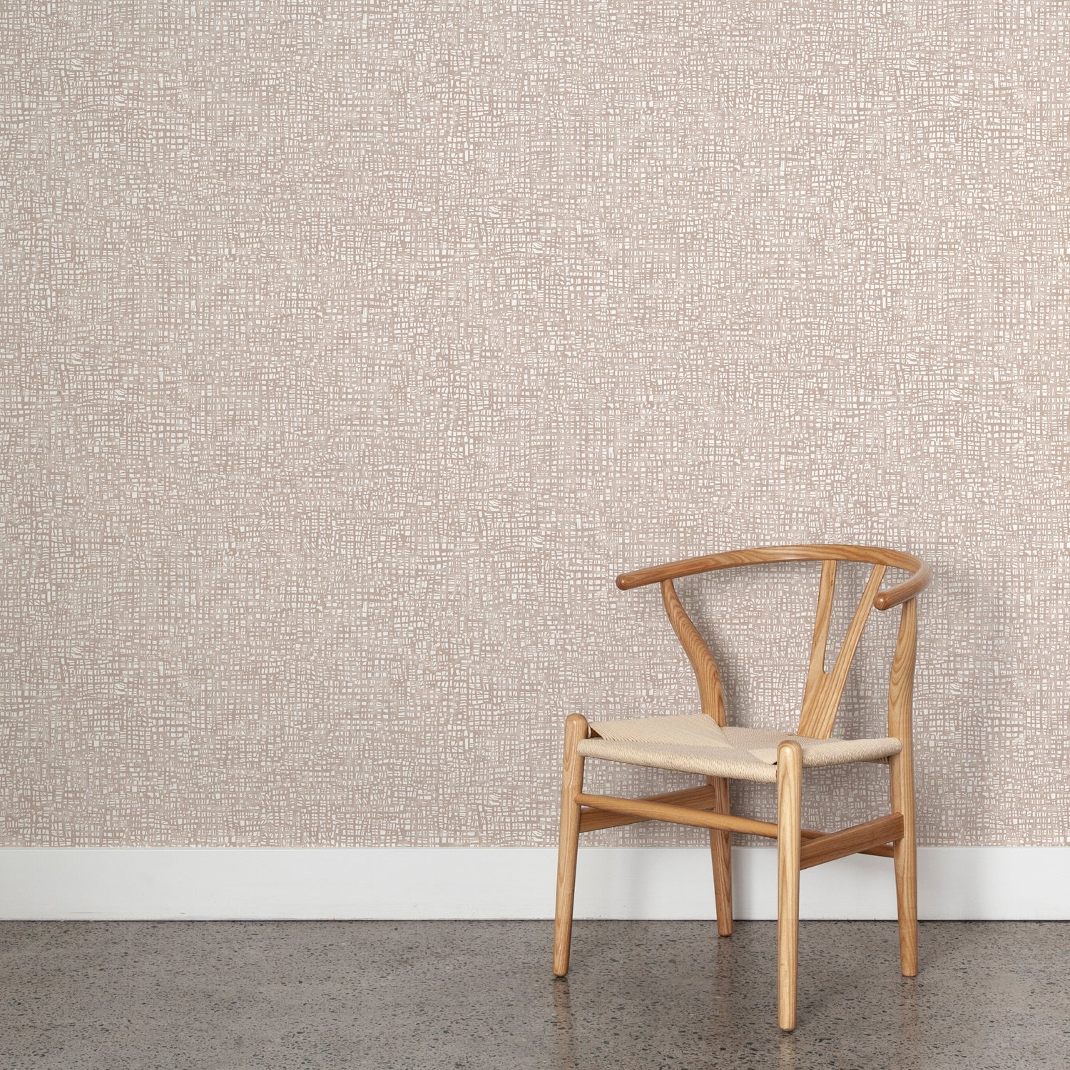 Angled wooden chair with woven seat on concrete floor against wallpaper in a textural wicker pattern in muted blush pink on a white field.