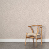 Angled wooden chair with woven seat on concrete floor against wallpaper in a textural wicker pattern in muted blush pink on a white field.
