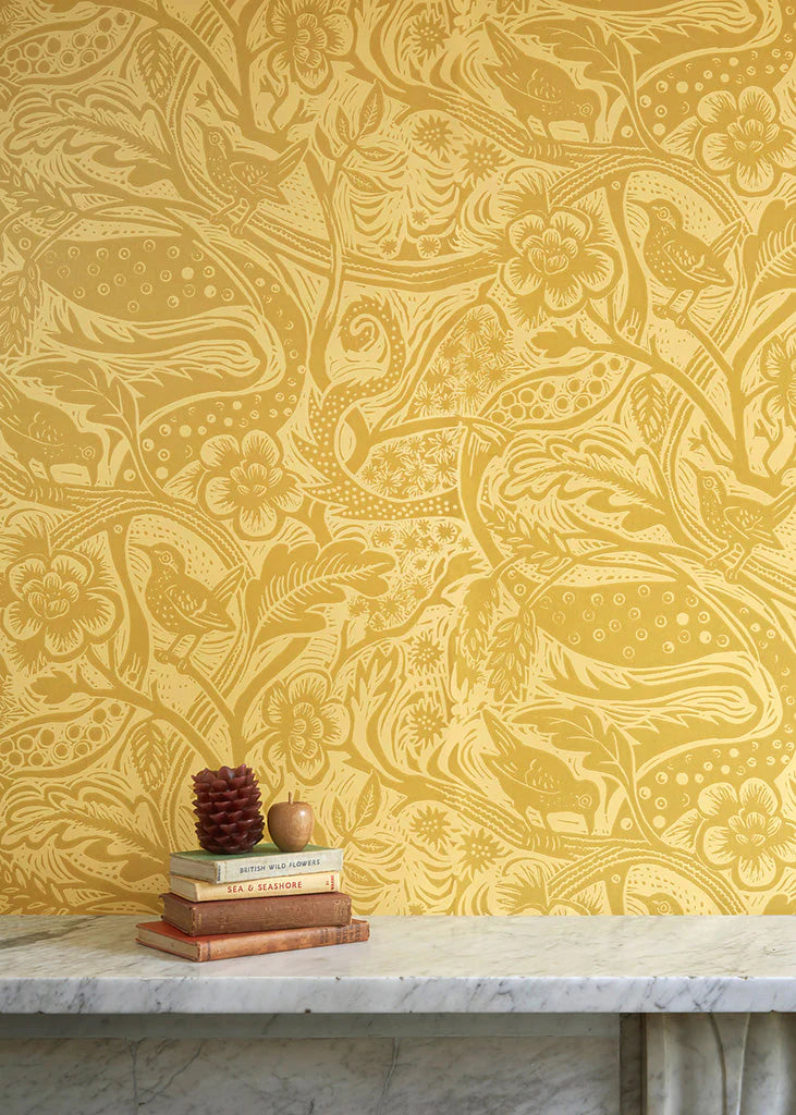 A stack of books stands in front of a wall papered in a playful wren and flower print in yellow and gold.