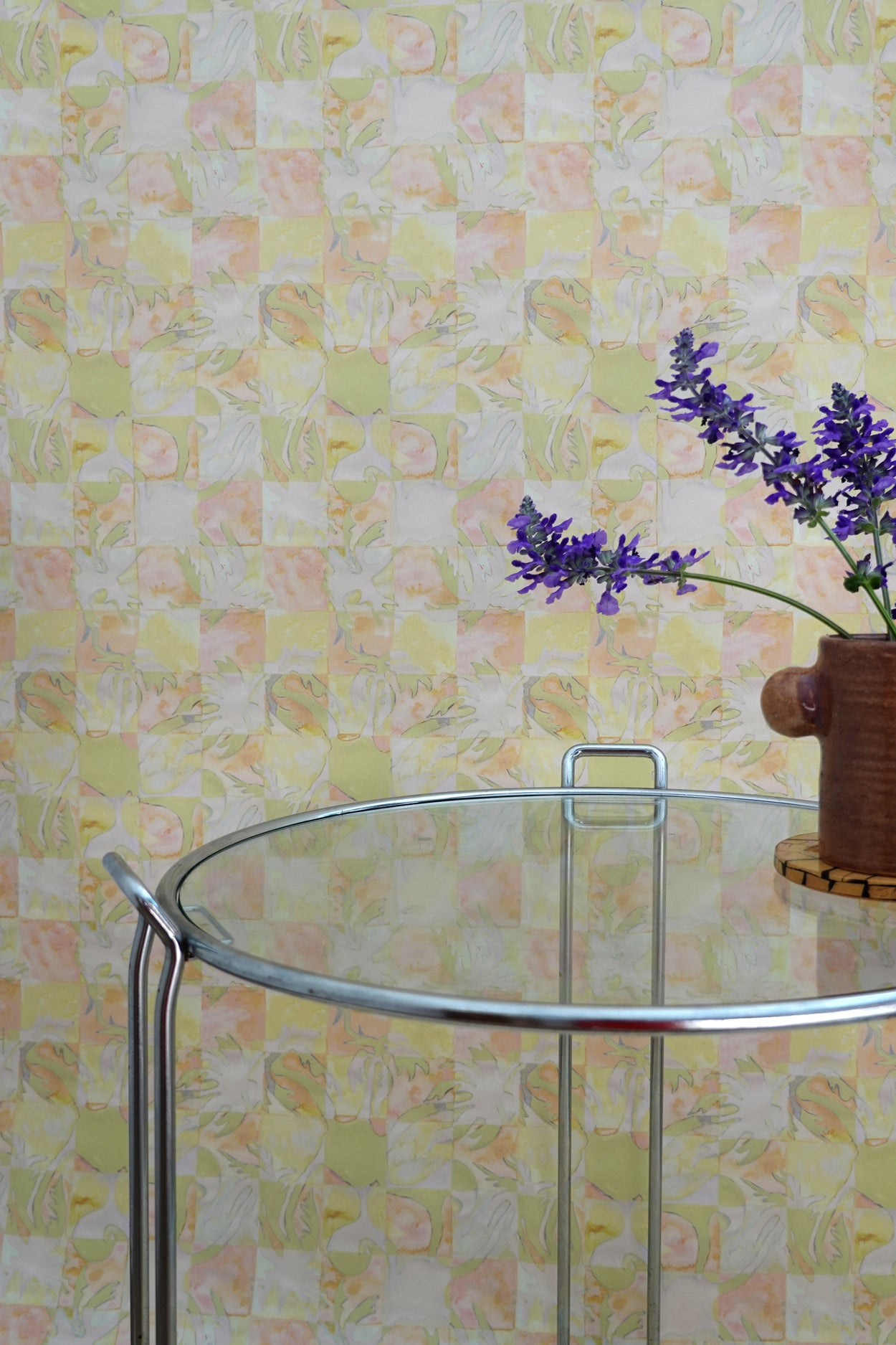 Glass table with a vase of purple flowers against a yellow tropical checkerboard patterned wall.