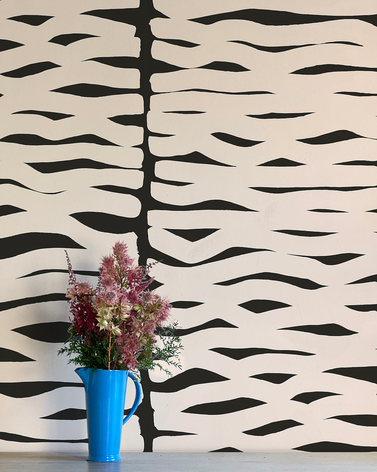 A vase of flowers stands in front of a wall papered in an abstract zigzag pattern in cream on a black field.