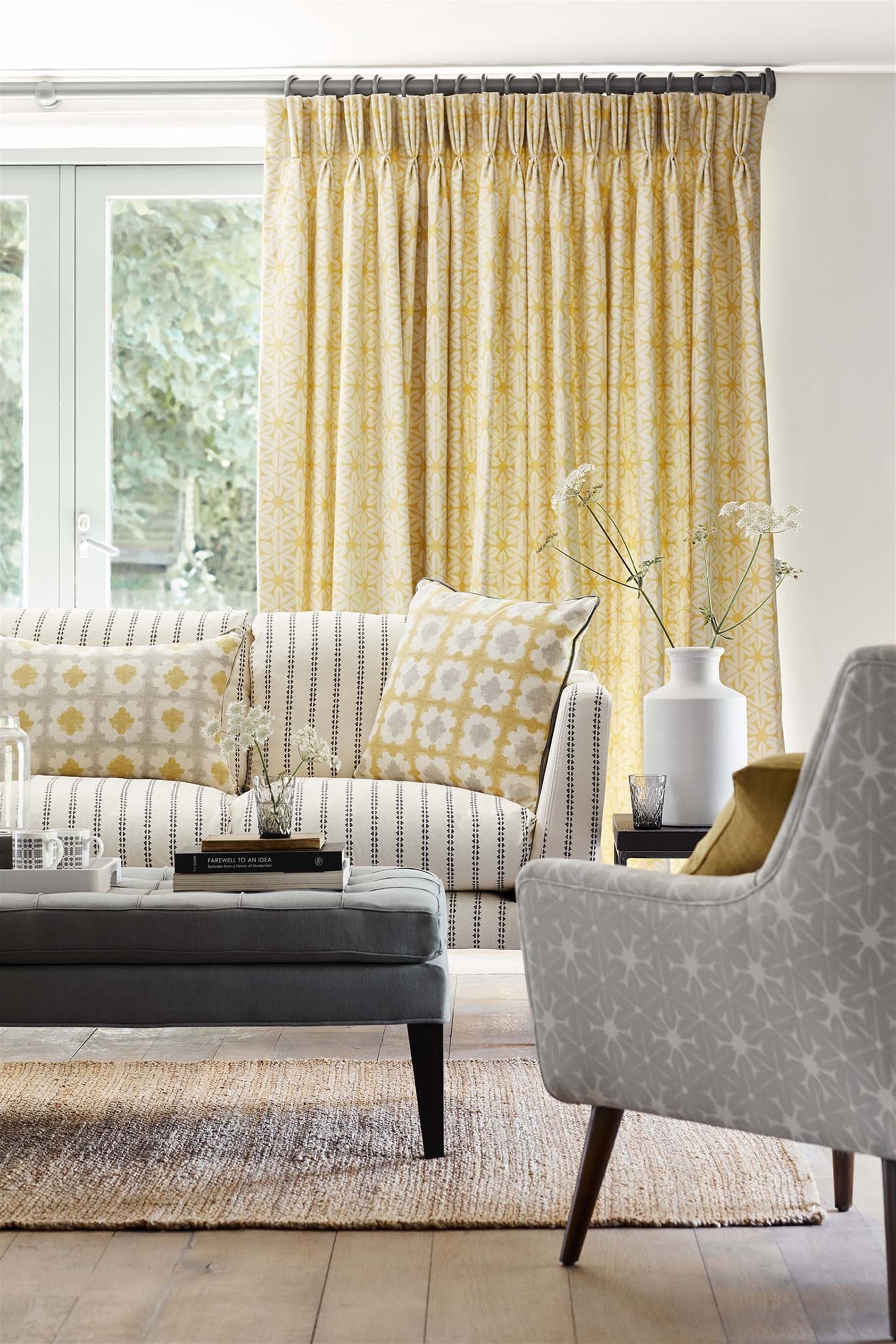 Living room scene with grey and cream upholsterd chair and coach with yellow throw pillows and a yellow printed curtain 