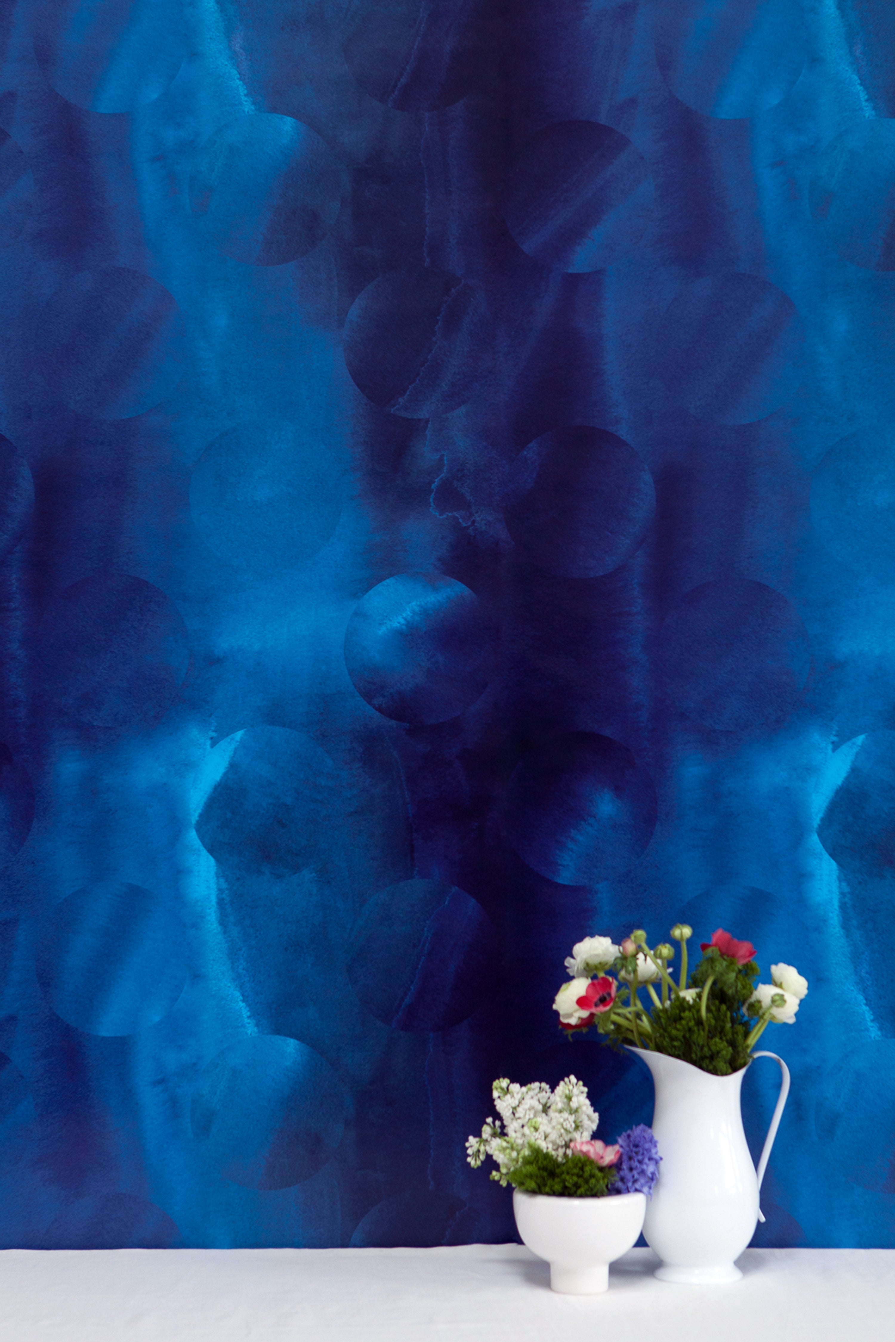 Two vases of flowers stand in front of a wall covered in an abstract curvilinear pattern in mottled blue, navy and black.