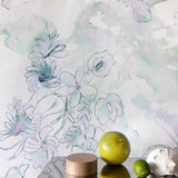 A table with fruit and knick knacks stands in front of a wall papered in a painterly floral print in green, purple and white.