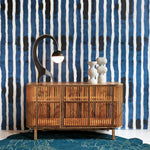 A mid-century modern wooden sideboard cabinet with a slatted bamboo front, styled with a black curved lamp and white ceramic figurines, set against a blue and white striped wallpaper background and a teal rug.