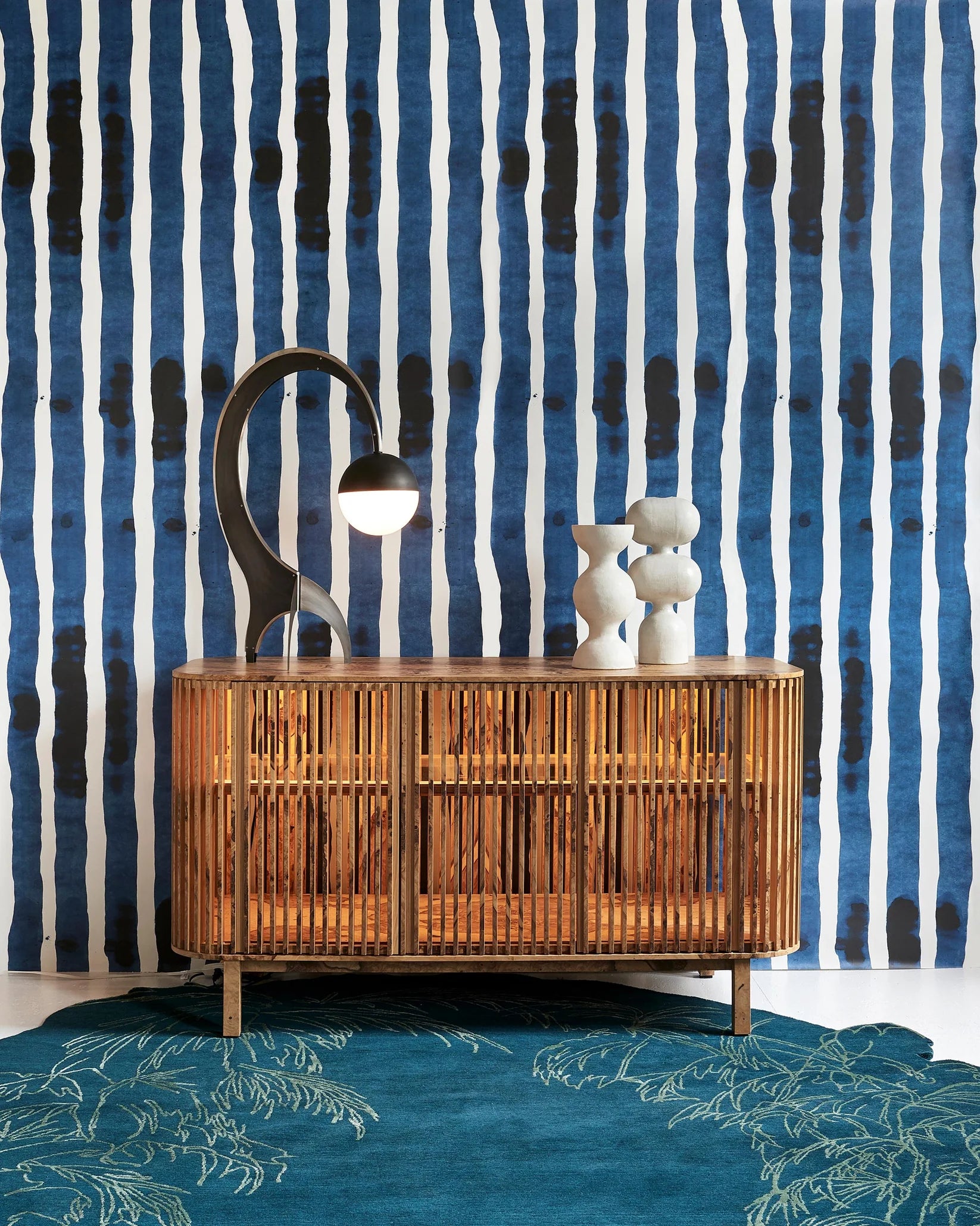 A mid-century modern wooden sideboard cabinet with a slatted bamboo front, styled with a black curved lamp and white ceramic figurines, set against a blue and white striped wallpaper background and a teal rug.
