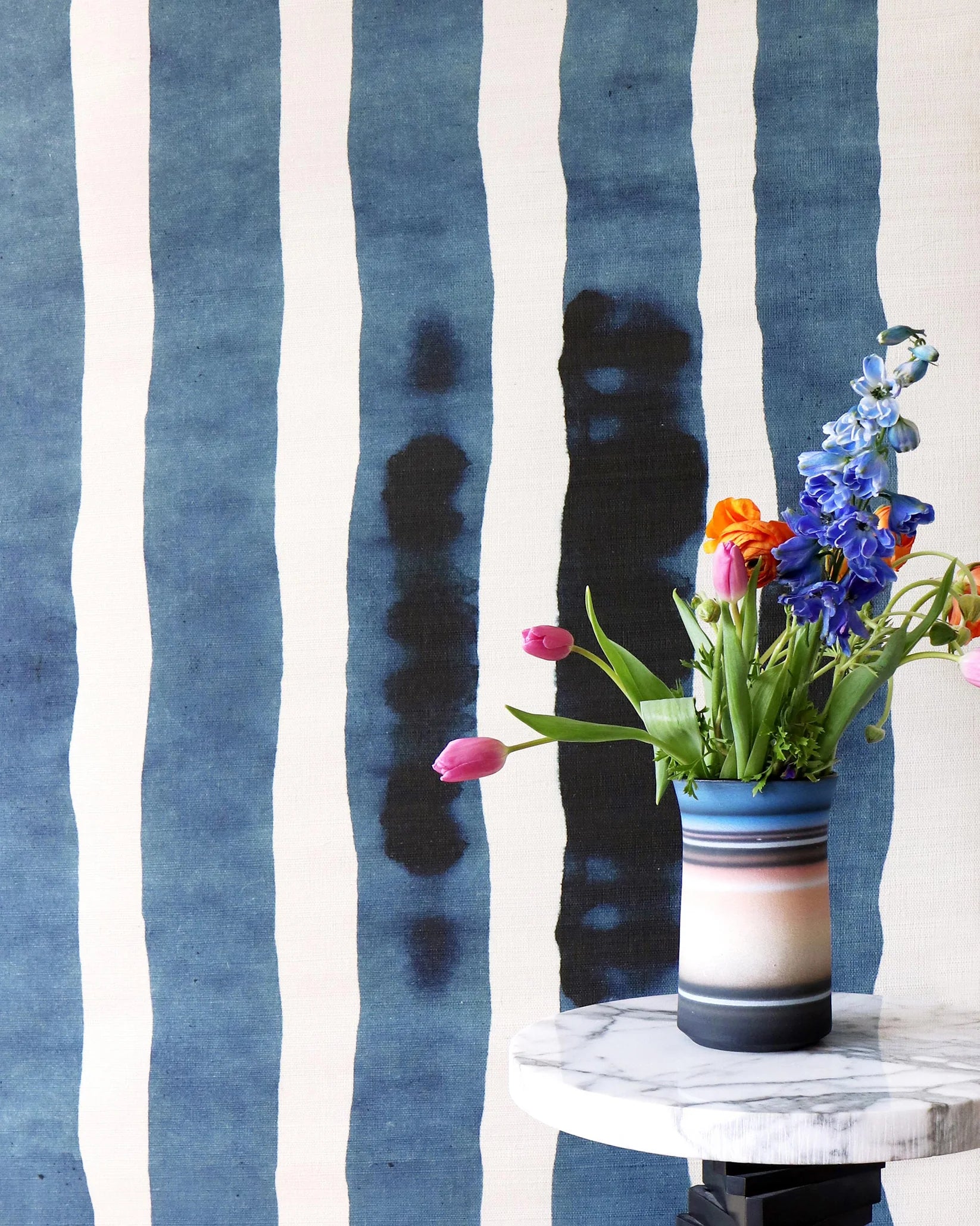 A vase of colorful tulips and flowers sits on a white marble side table against a blue and white watercolor stripe wallpaper.