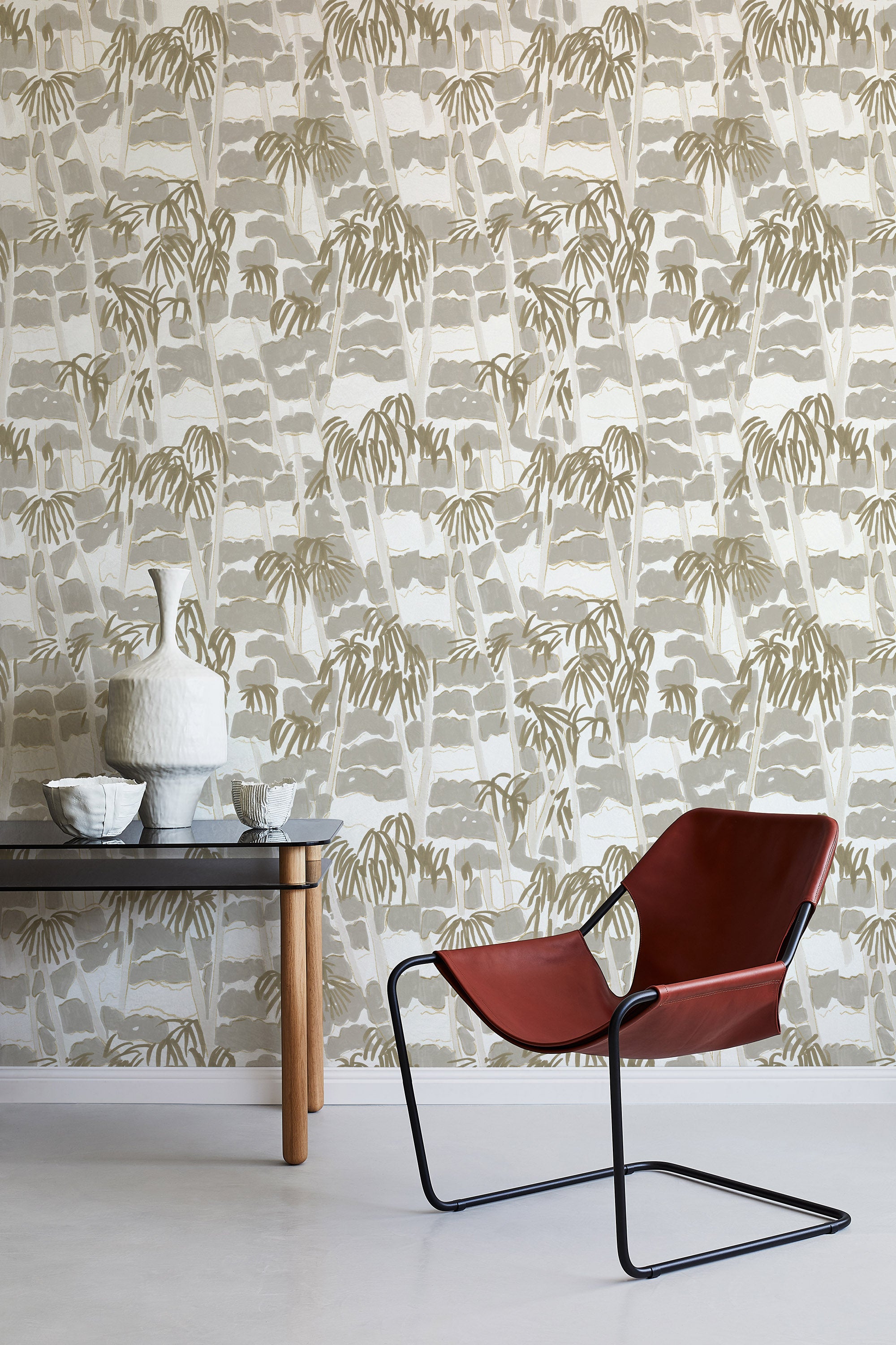 A modernist armchair stands in front of a wall papered in a painterly palm tree print in shades of gray, gold and white.