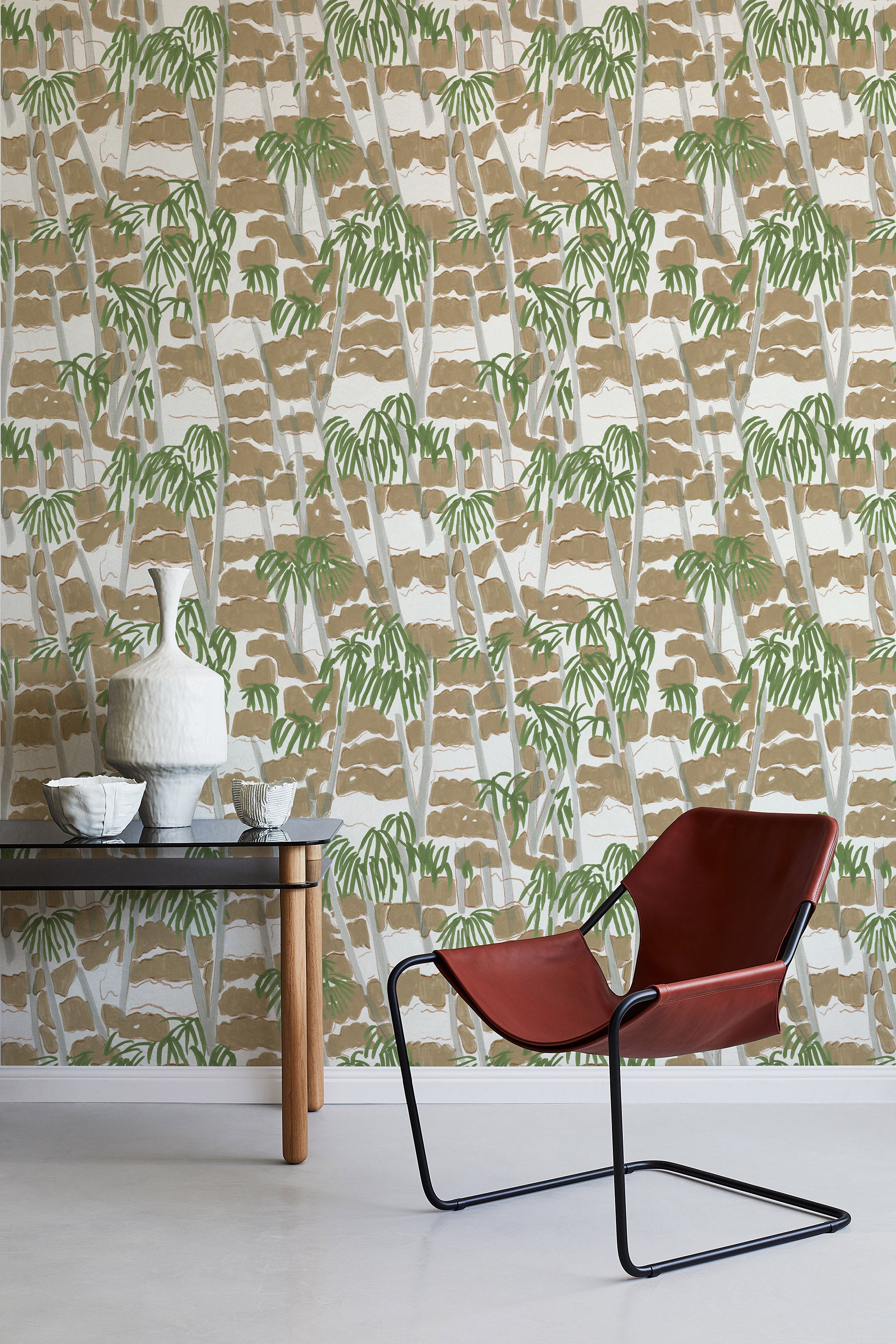 A modernist armchair stands in front of a wall papered in a painterly palm tree print in shades of green, gray and brown.