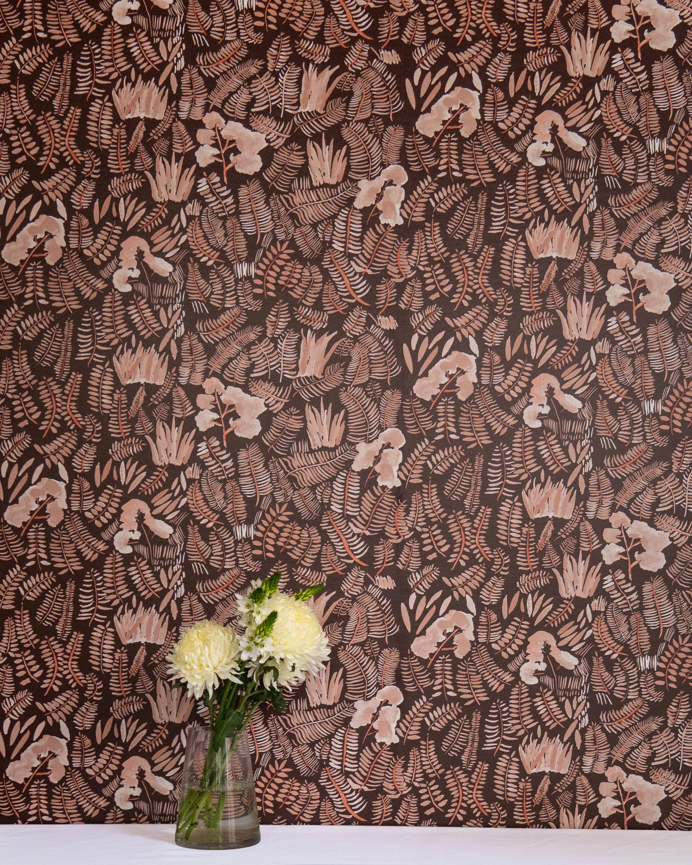 A vase of flowers stands in front of a wall papered in a playful leaf print in shades of orange and coral on a brown field.