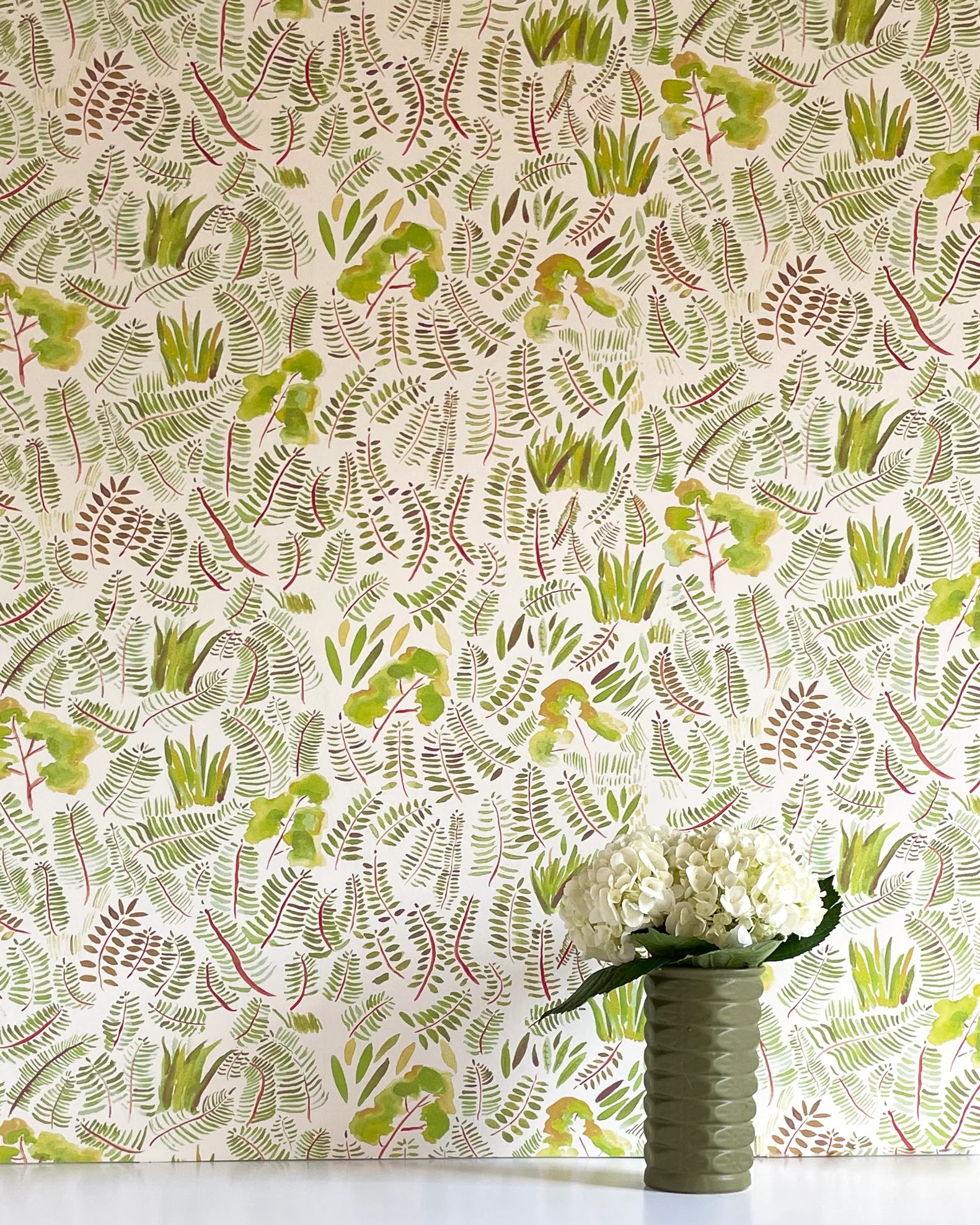 A vase of flowers stands in front of a wall papered in a playful leaf print in shades of green and red on a cream field.