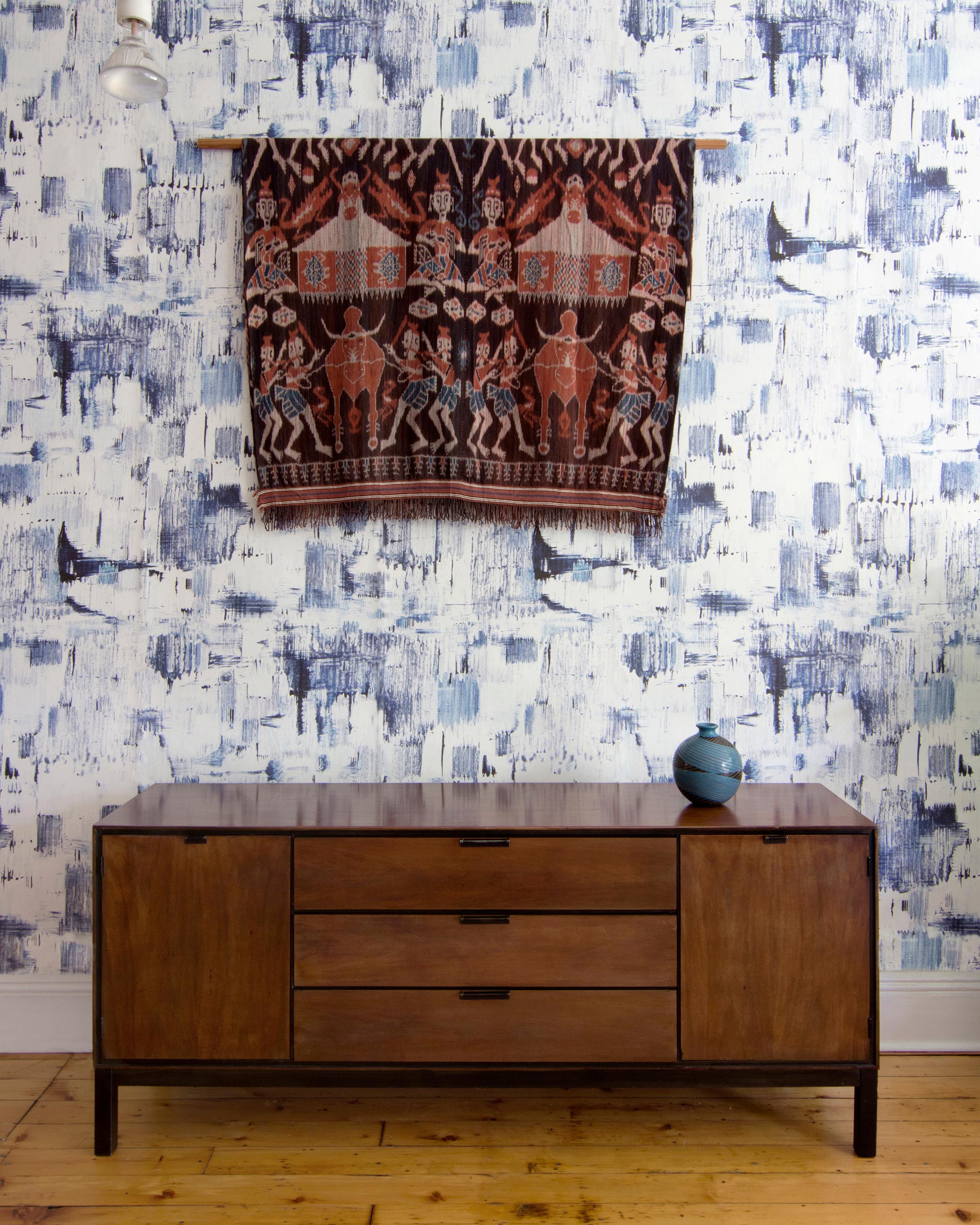 A credenza and tapestry stand in front of a wall papered in a watercolor textural print in navy, blue and white.