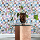 An end table with a large potted plant stands in front of a wall papered in a chess board print in a rainbow of shades.