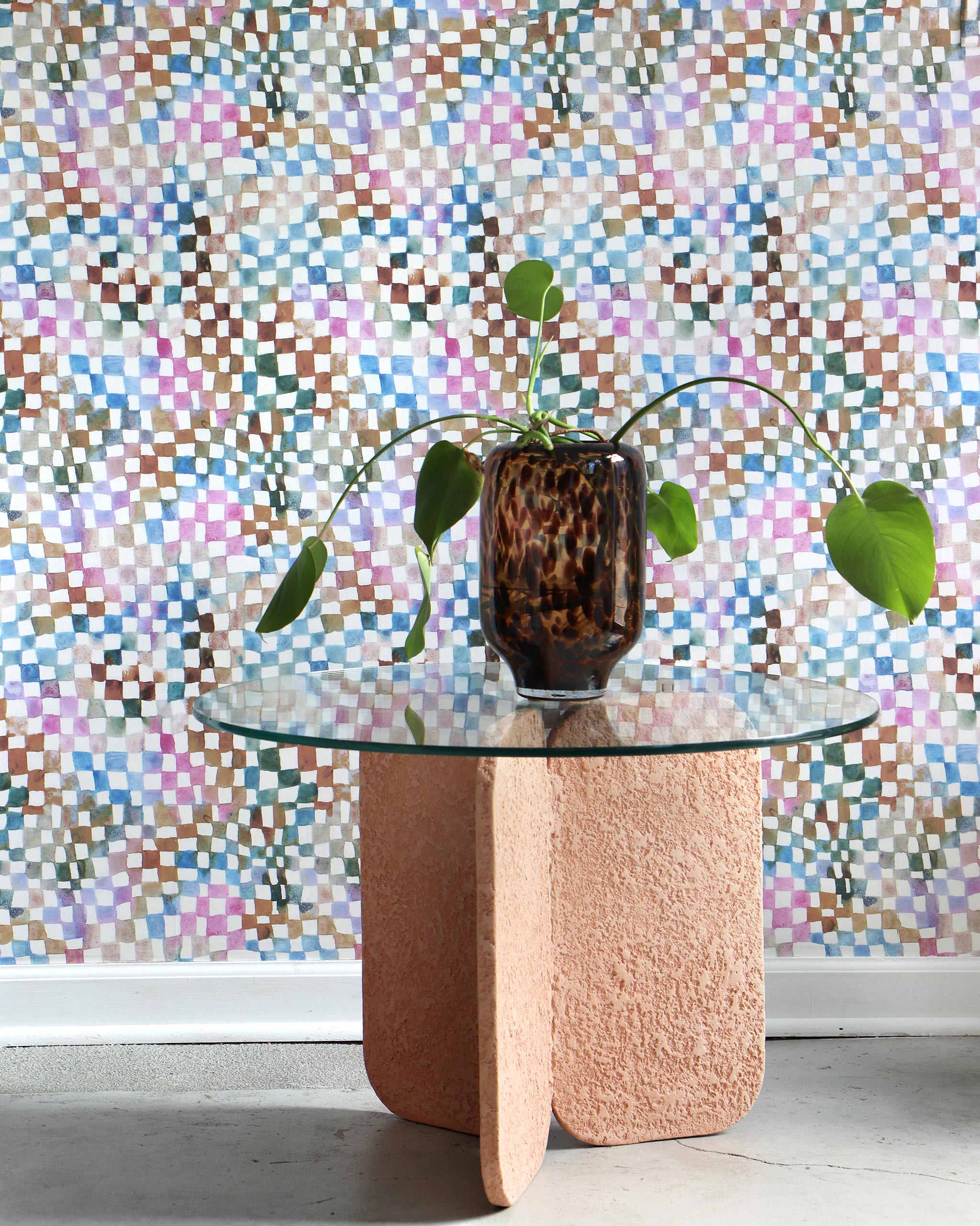 An end table with a large potted plant stands in front of a wall papered in a chess board print in a rainbow of shades.