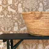 A basket sits on a bench in front of a wall papered in a lion and plant print in white on a mottled cream field.