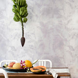 A table covered in fruit stands in front of a wall papered in a painterly palm tree print in shades of gray and cream.