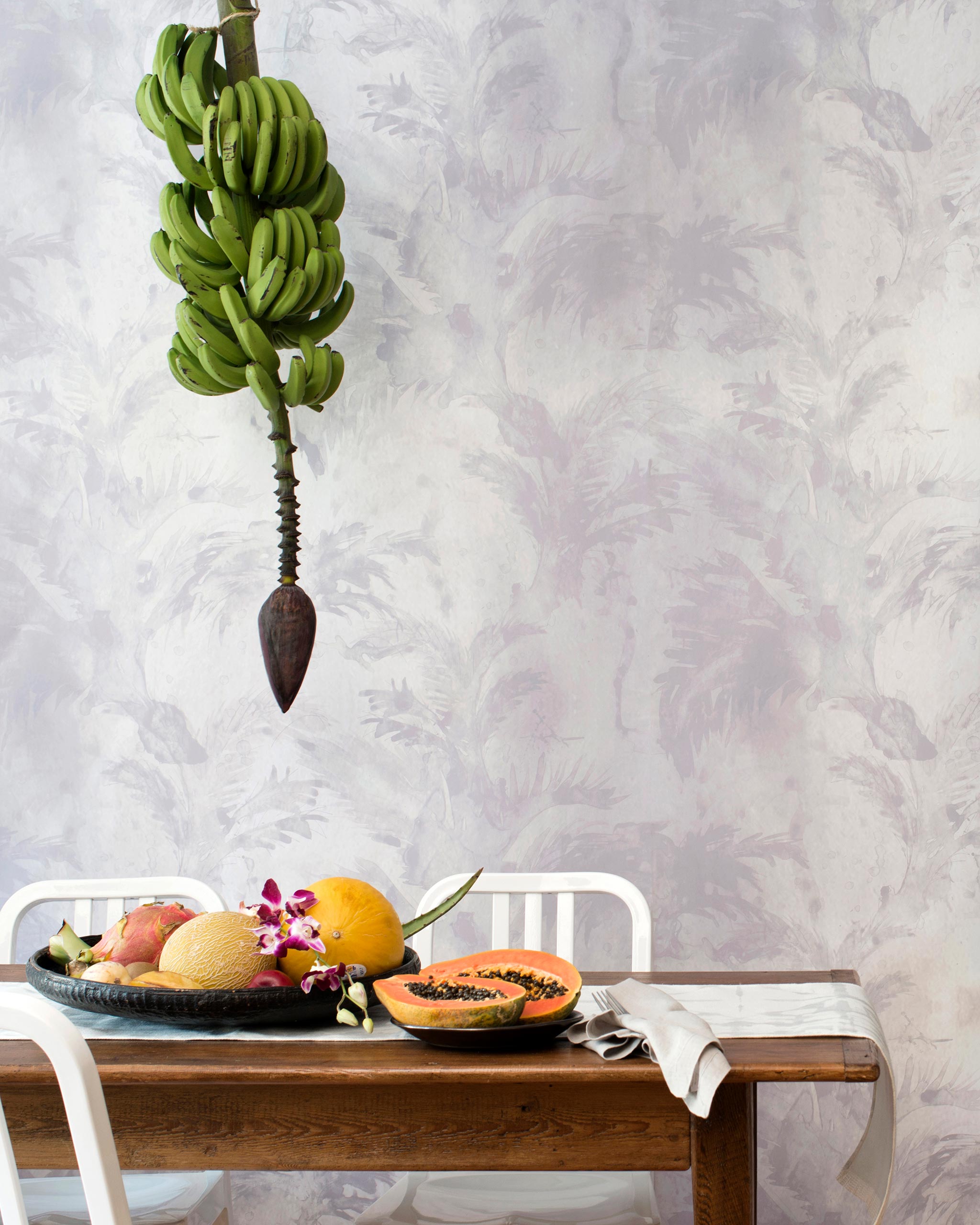 A table covered in fruit stands in front of a wall papered in a painterly palm tree print in shades of gray and cream.