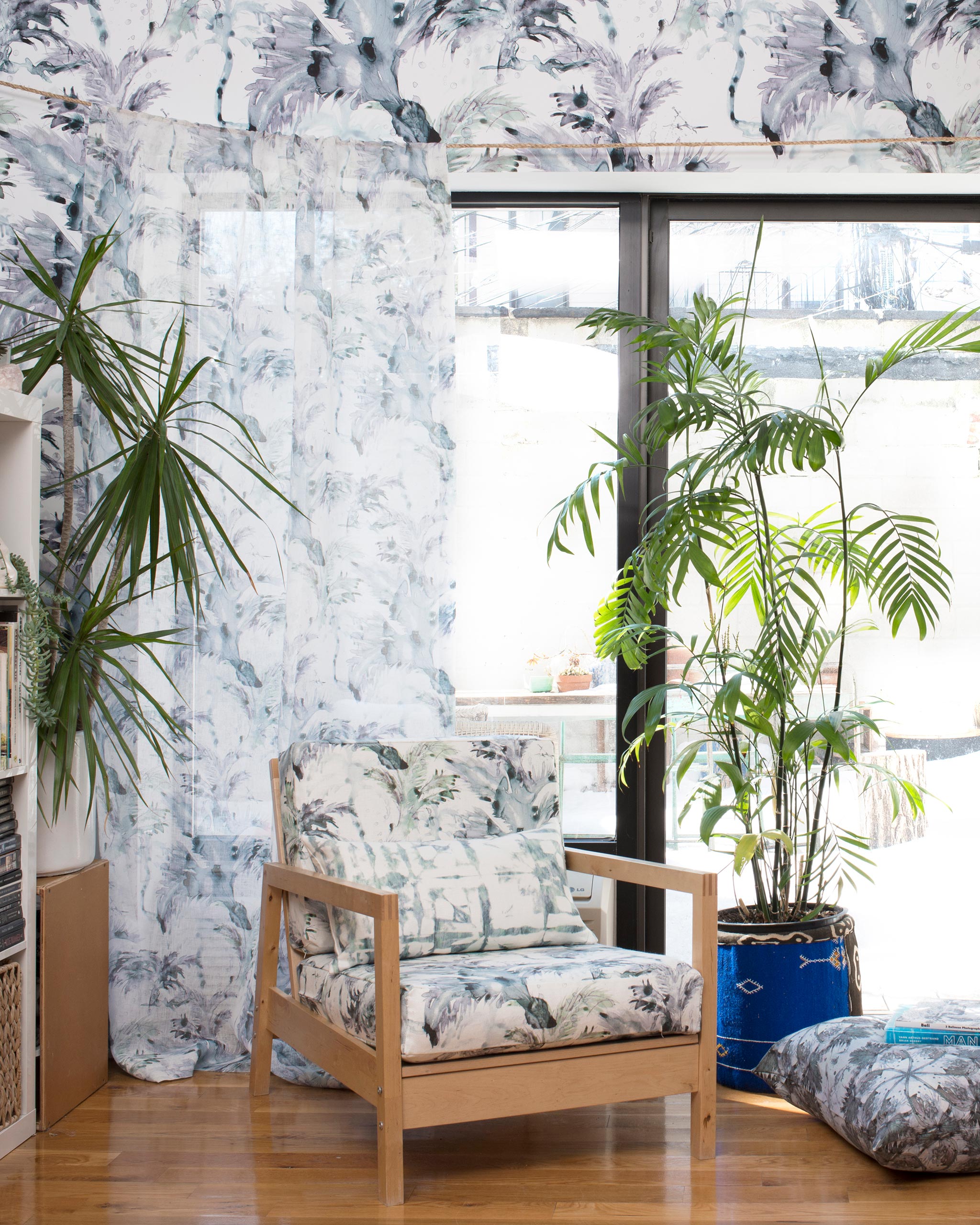 A plant-filled living space with an armchair upholstered in a painterly palm leaf print in turquoise, gray and white.