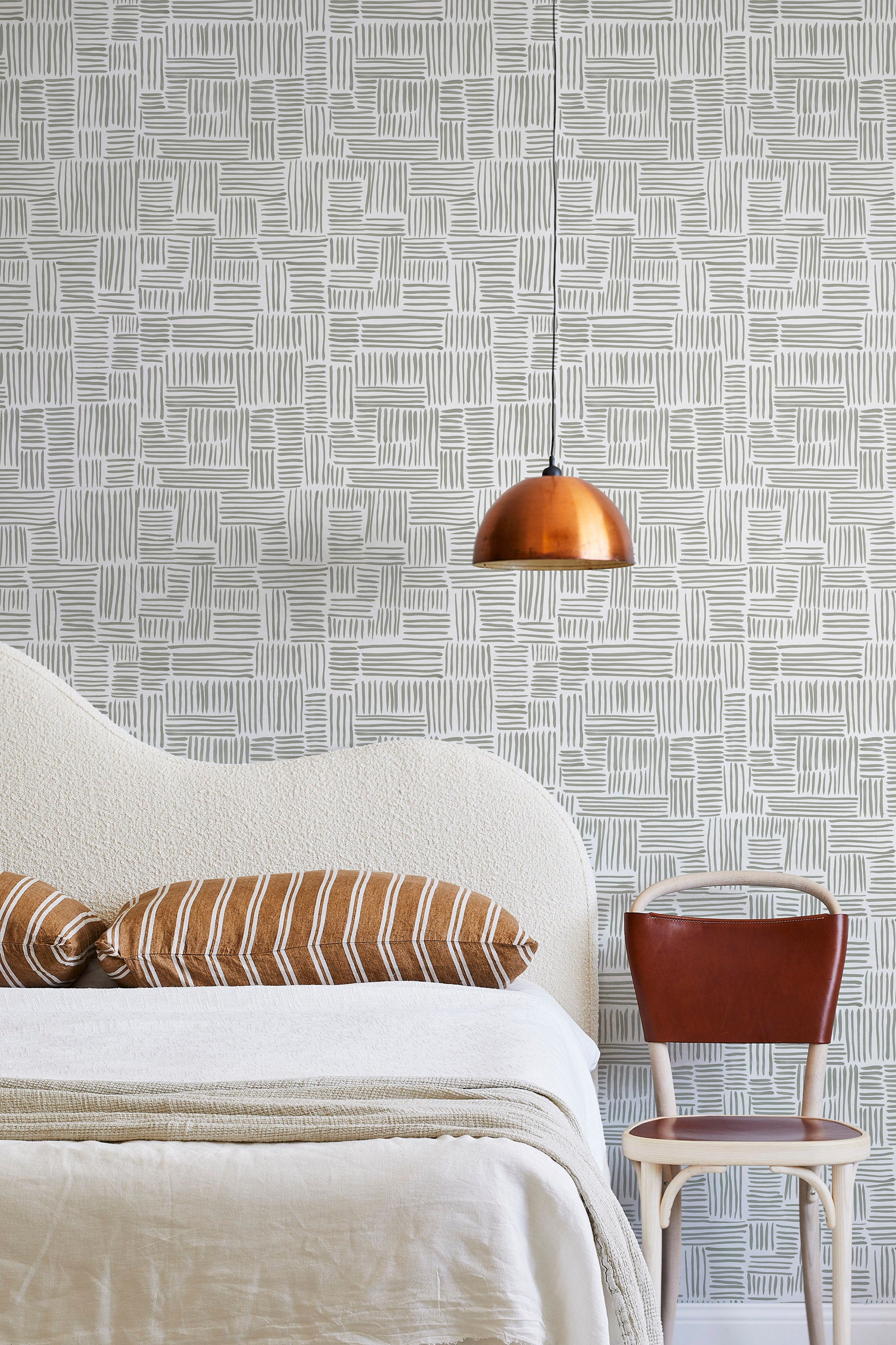 A modernist bed, hanging lamp and chair stand in front of a wall papered in a directional dash pattern in green-gray and white.