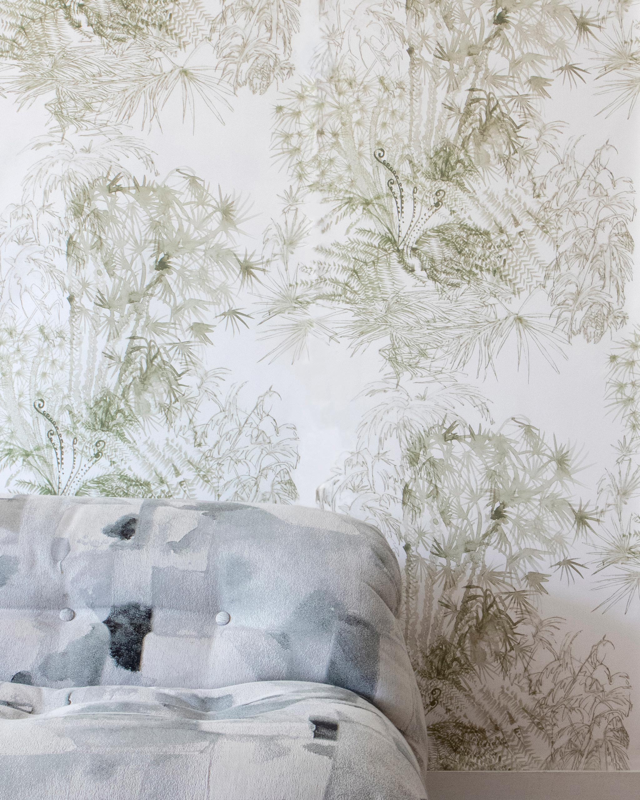 A bed stands in front of a wall papered in a painterly palm tree print in olive on a cream field.