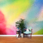 A chair, plant and cat stand in front of a wall covered in an ombré paint splatter print in pink, yellow, blue and green.