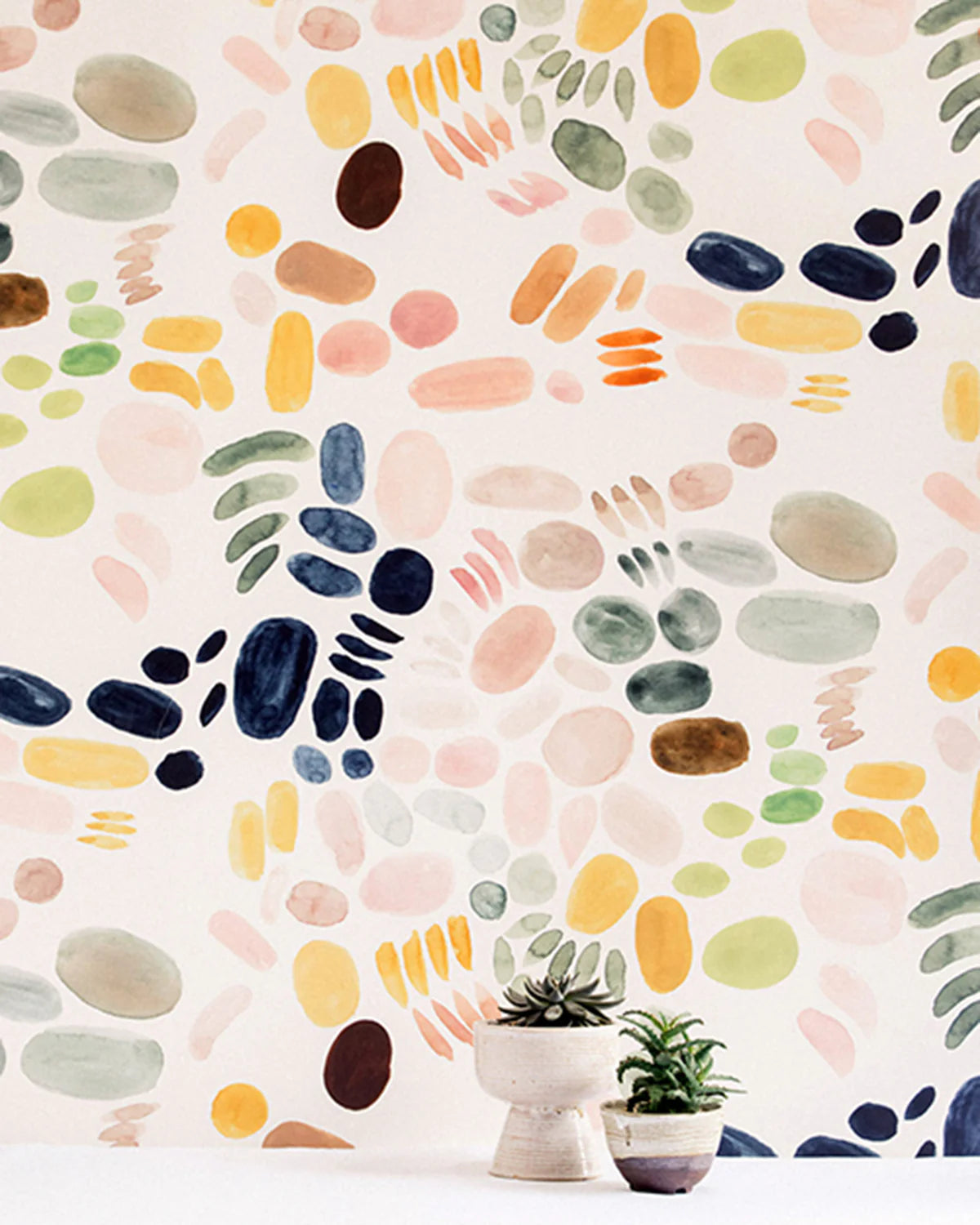 A vase of flowers stands in front of a wall covered in a playful paint blotch print in green, pink and yellow on a white field.