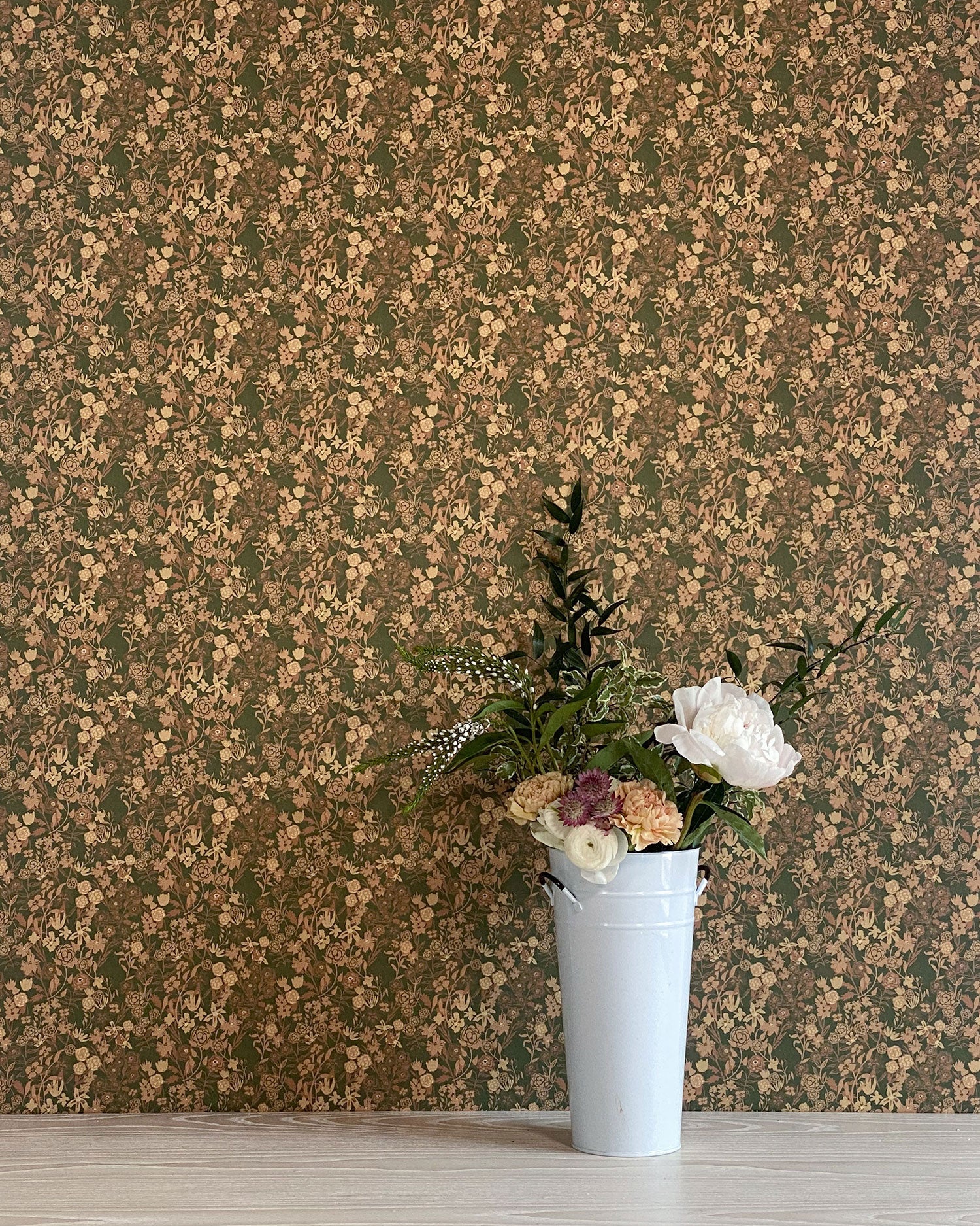 A vase of flowers stands in front of a wall papered in a dense small-scale floral print in shades of brown, tan and green.