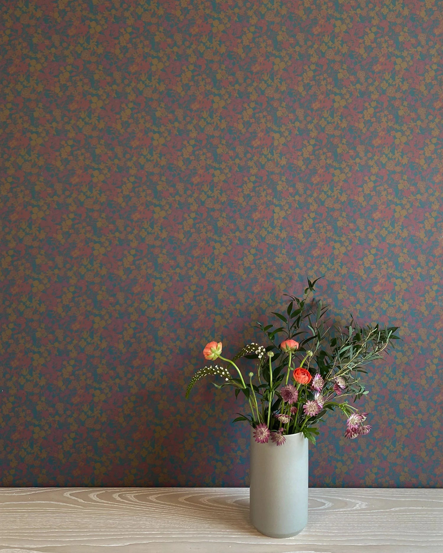 A vase of flowers stands in front of a wall papered in a dense small-scale floral print in shades of purple, gold and navy.