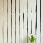 A vase of flowers stands in front of a wall covered in a playful irregular stripe print in black on a cream field.