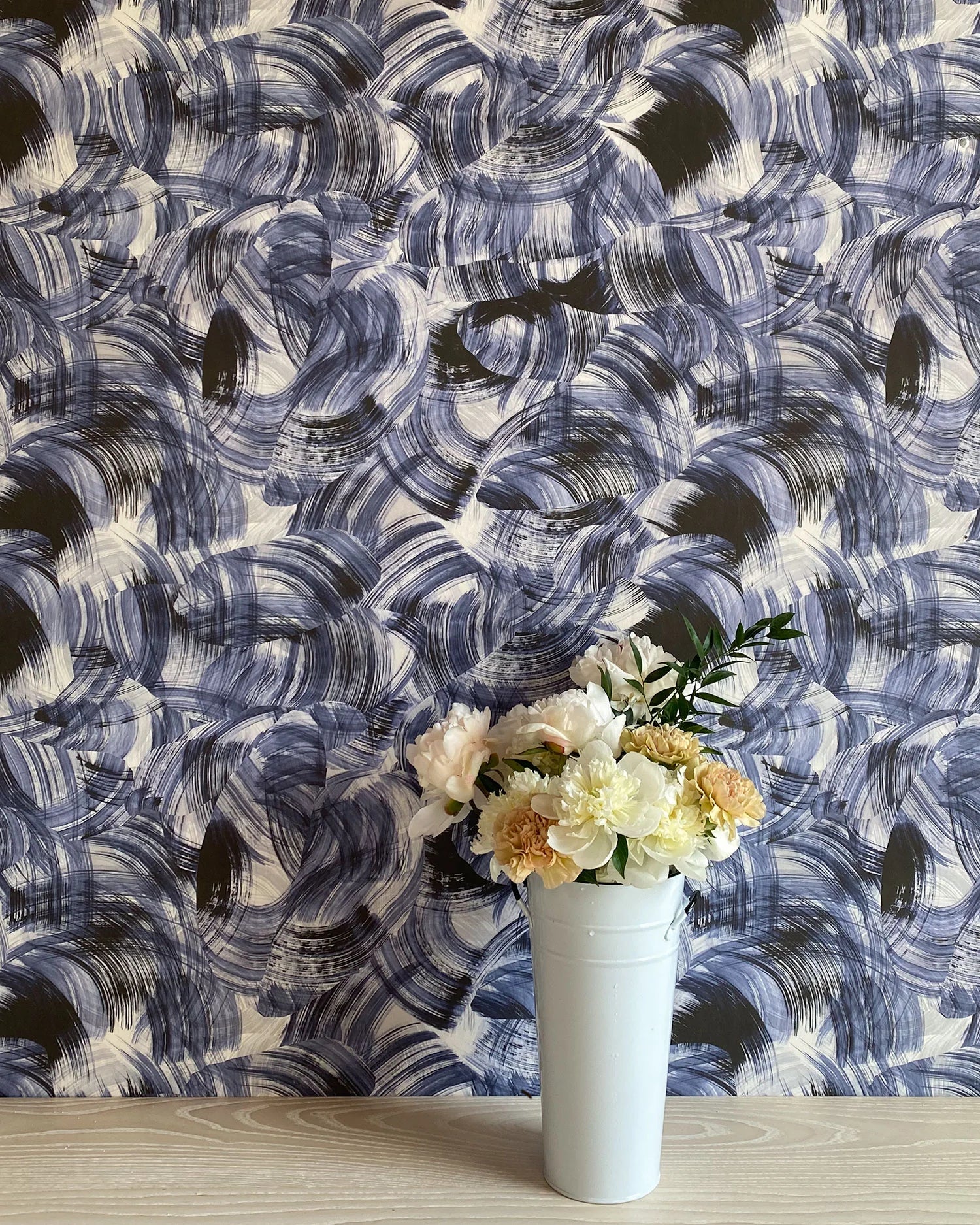 A vase of flowers stands in front of a wall papered in an abstract paint swatch print in black and navy on a cream field.