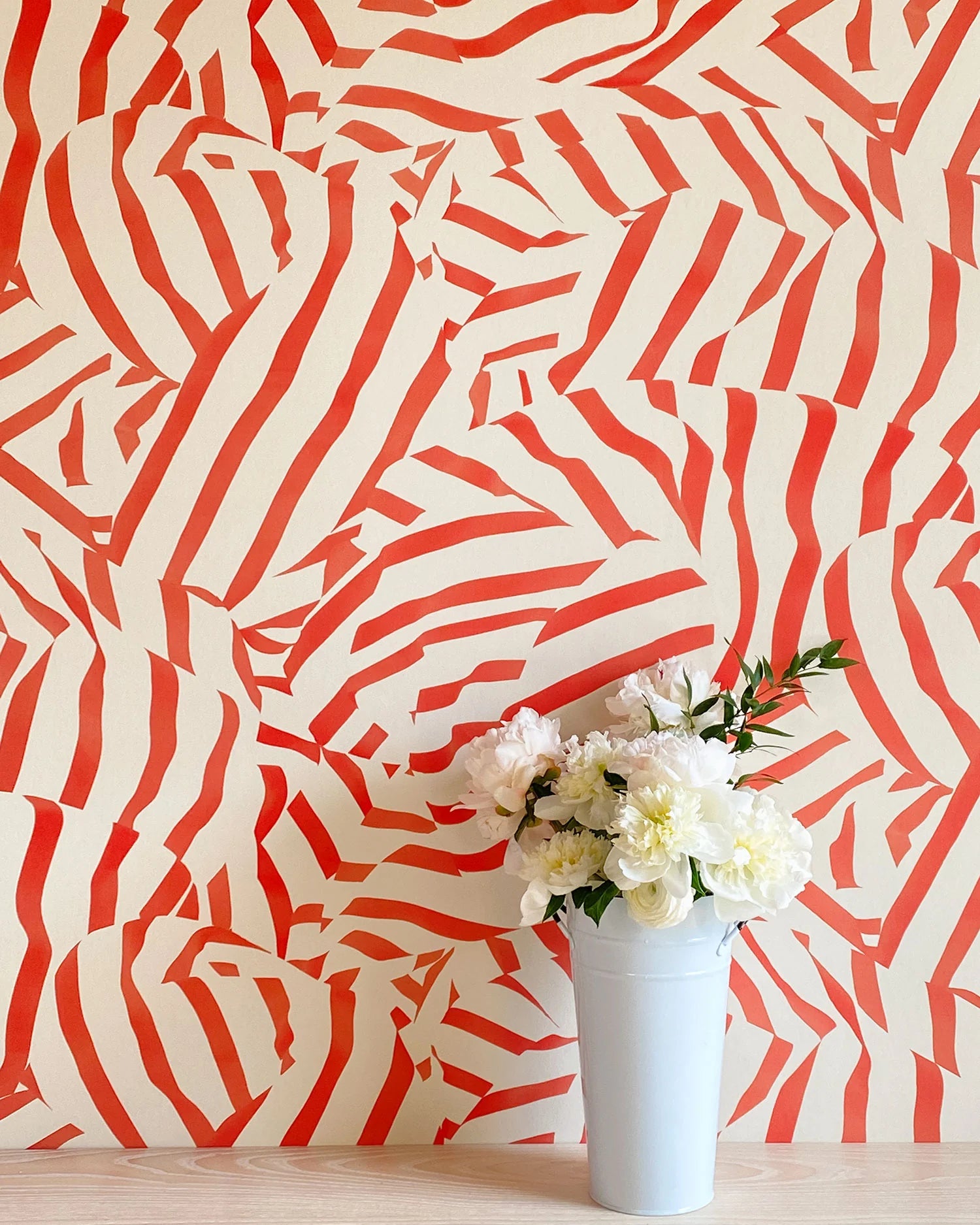 A vase of flowers stands in front of a wall papered in a playful broken stripe print in red on a cream field.