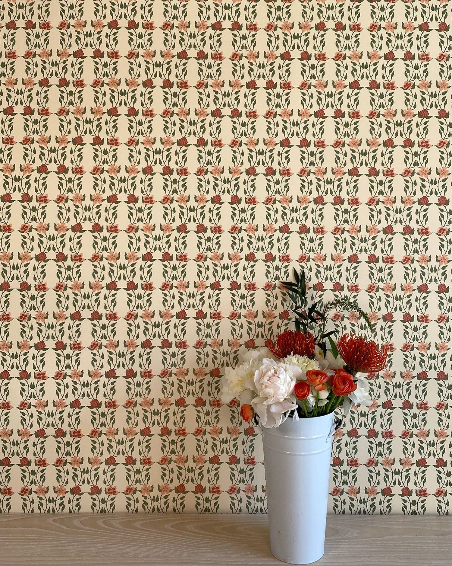 A vase of flowers stands in front of a wall papered in a linear floral print in red, orange and green on a cream field.