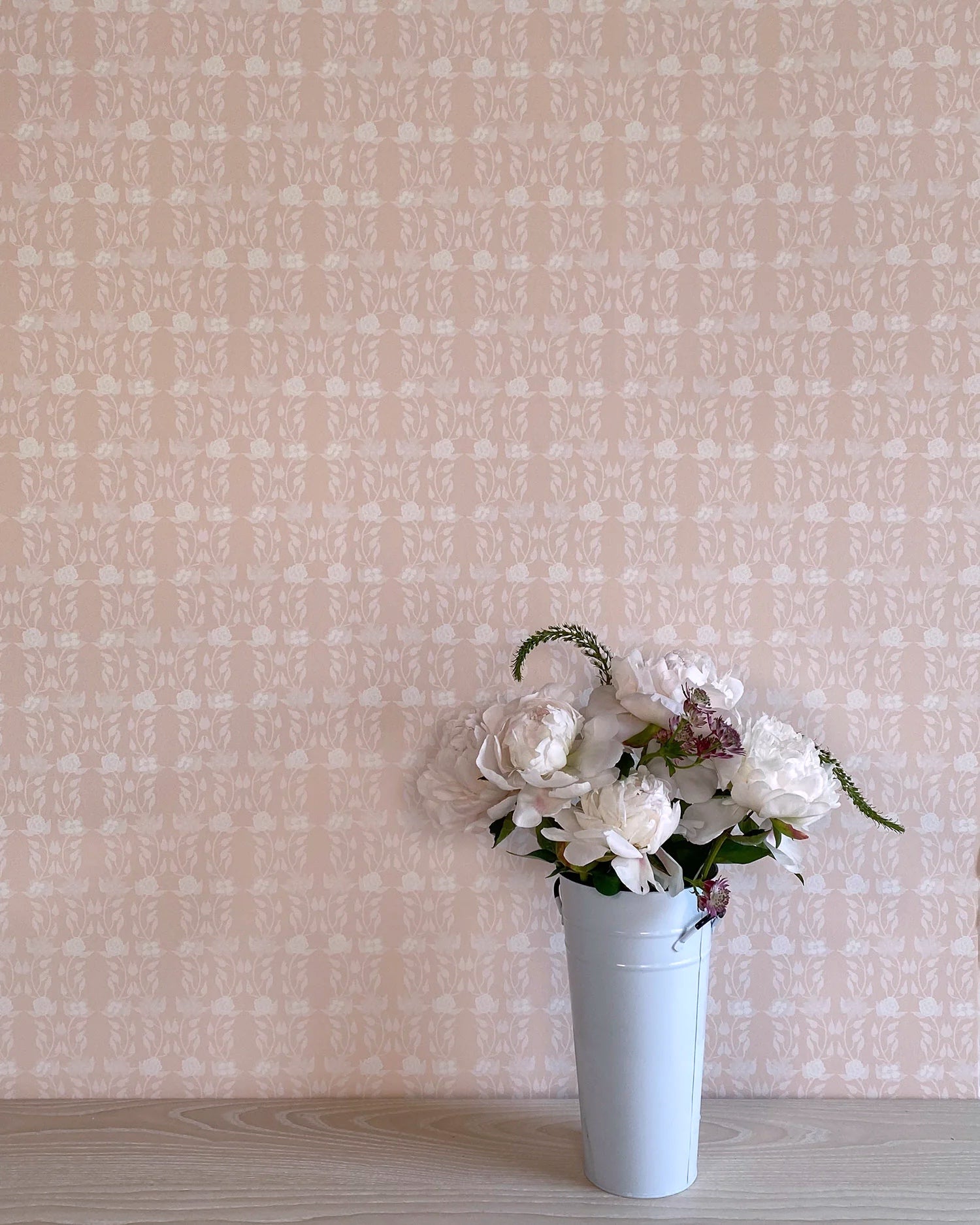 A vase of flowers stands in front of a wall papered in a linear floral print in white and cream on a light pink field.