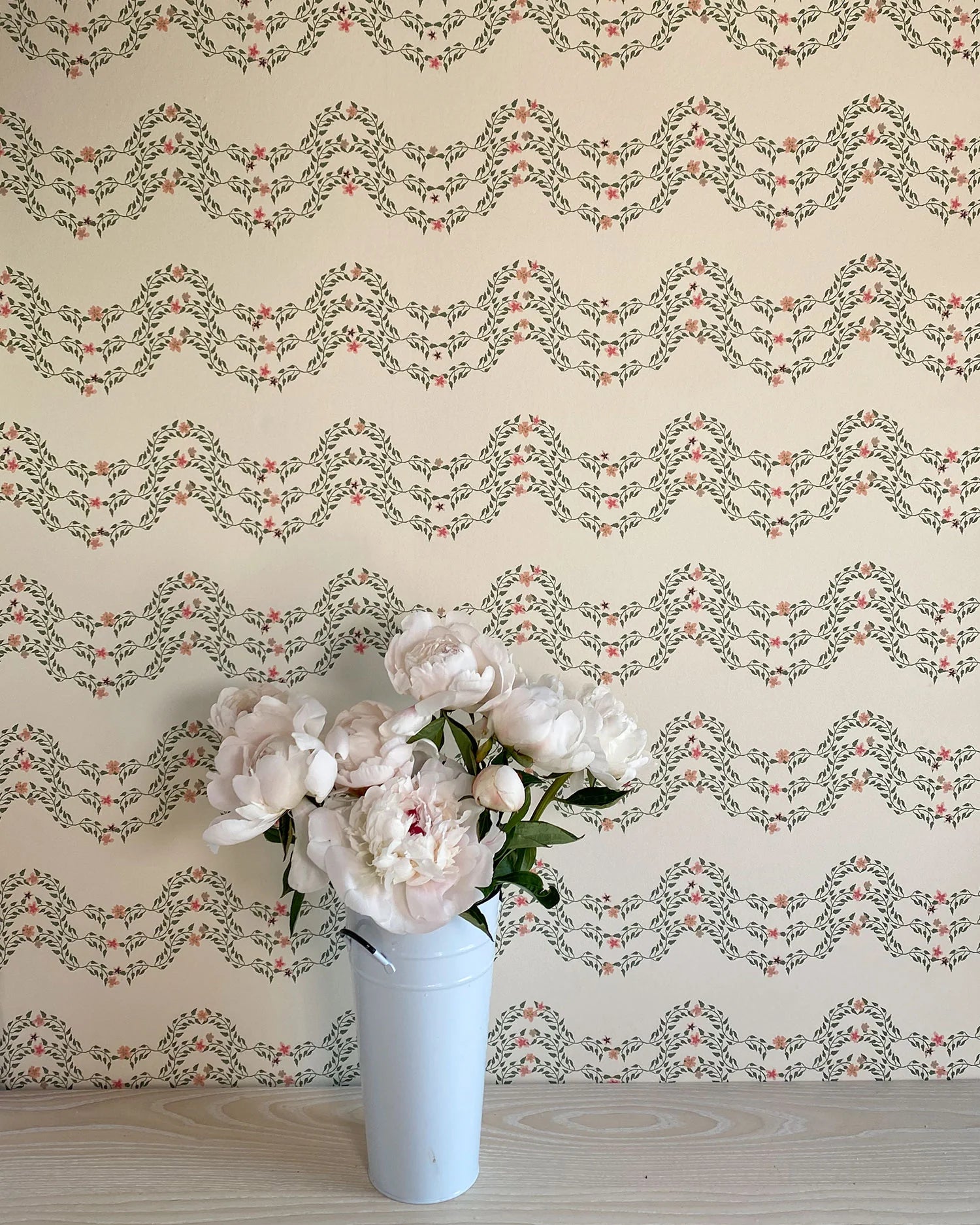 A vase of flowers stands in front of a wall covered in a curving floral stripe print in shades of orange and green on a cream field.