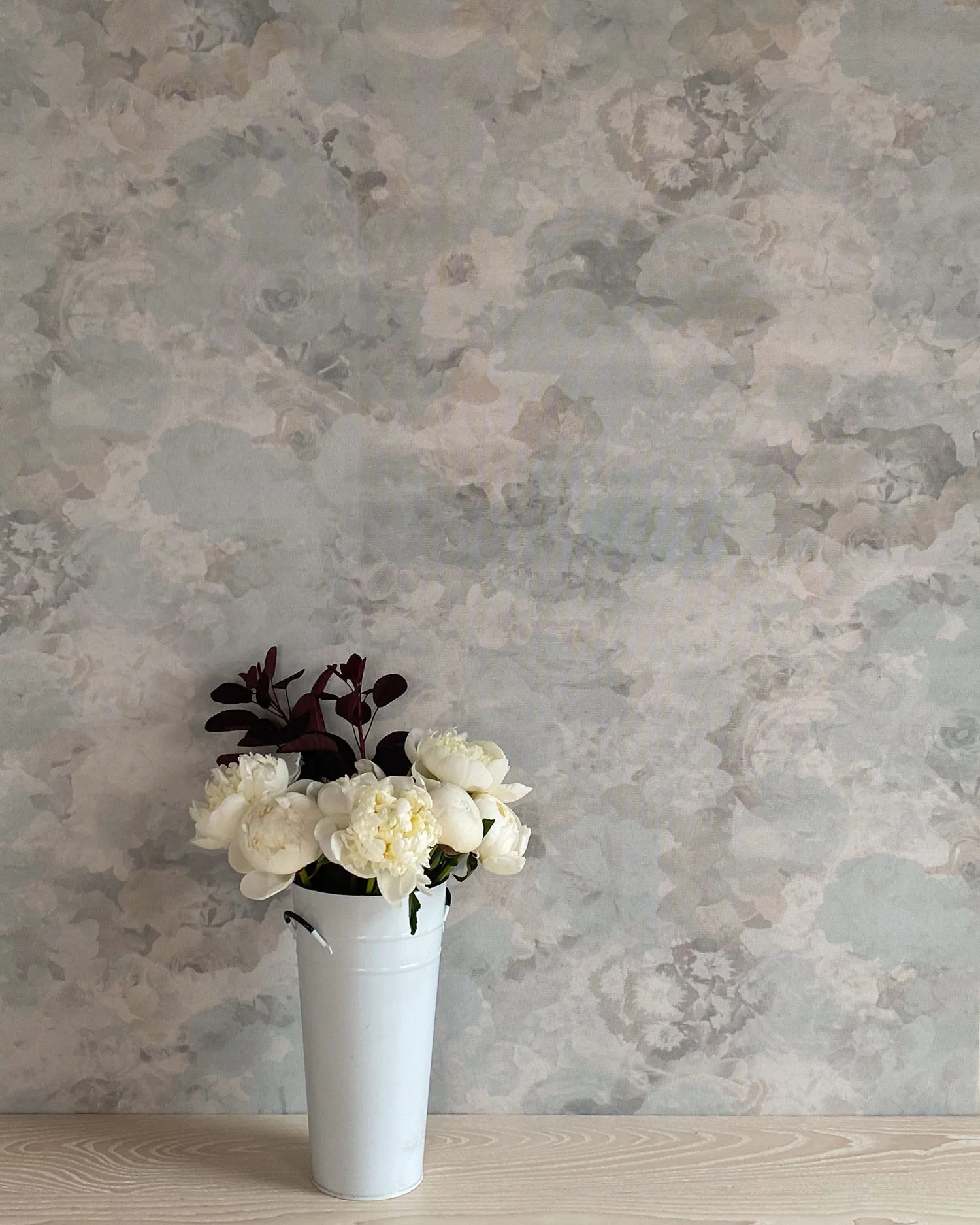 A vase of flowers stands in front of a wall covered in a dense photorealistic floral print in gray.