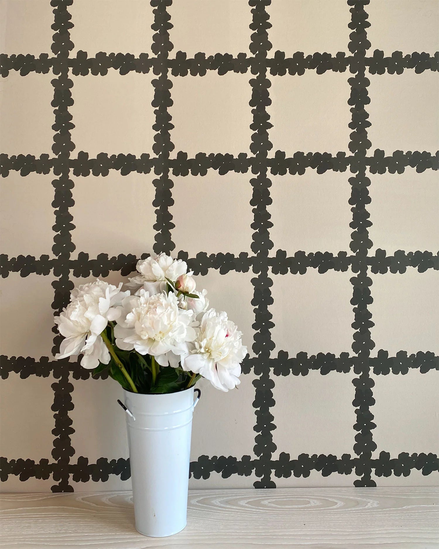 A vase of flowers stands in front of a wall covered in a floral grid print in brown on a white field.