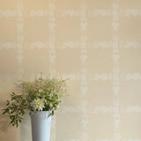A vase of flowers stands in front of a wall covered in a floral grid print in cream on a tan field.