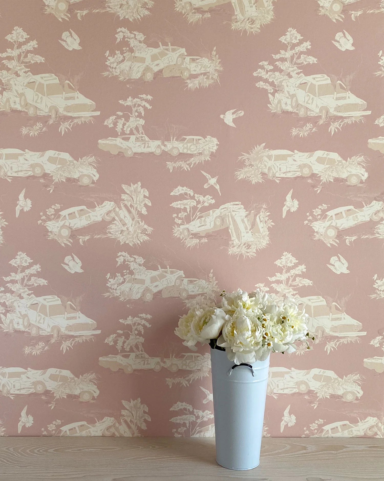 A vase of flowers stands in front of a wall covered in a playful illustrated car crash print in cream, white and mauve.