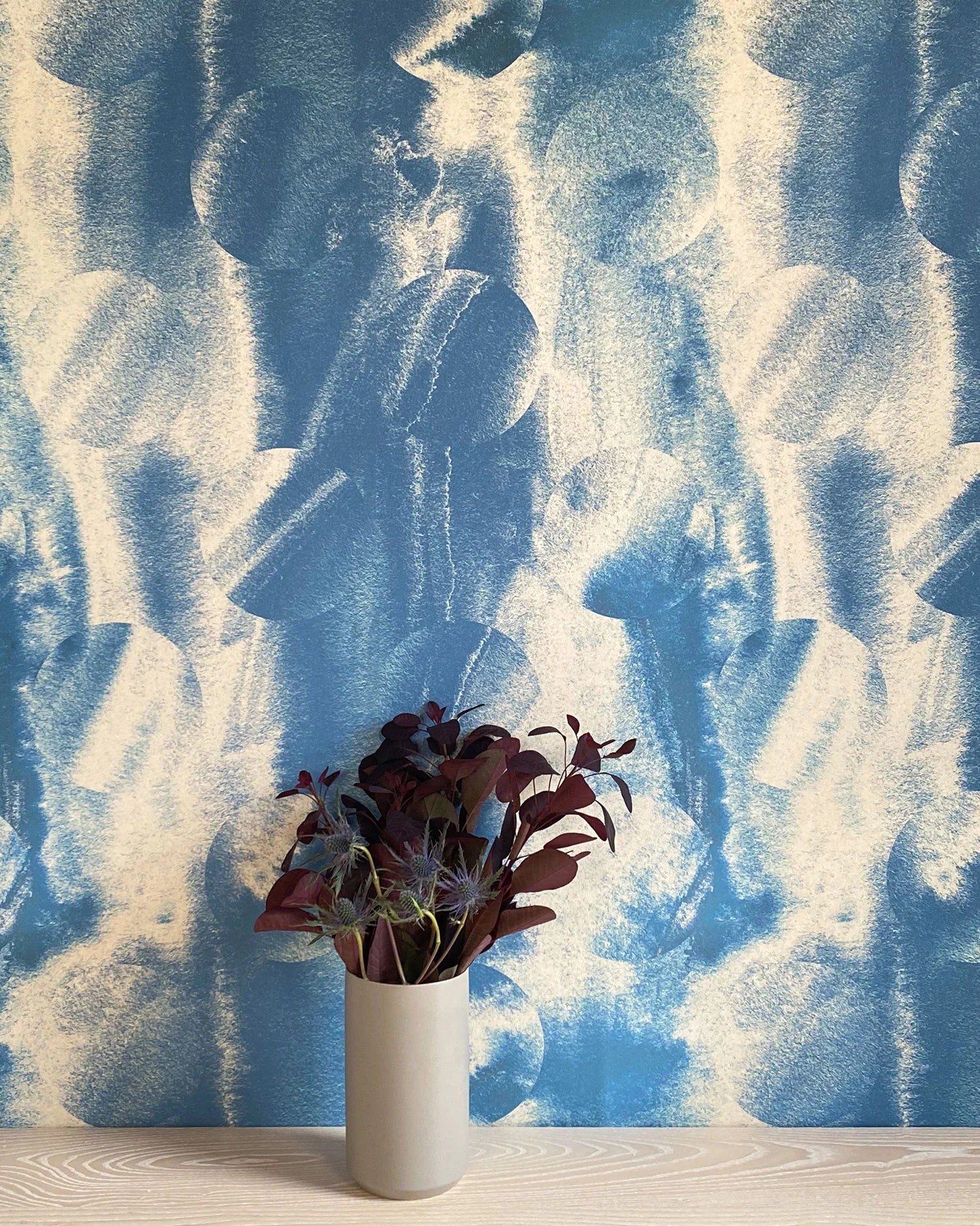 A vase of flowers stands in front of a wall papered in an abstract curvilinear pattern in mottled blue and tan.