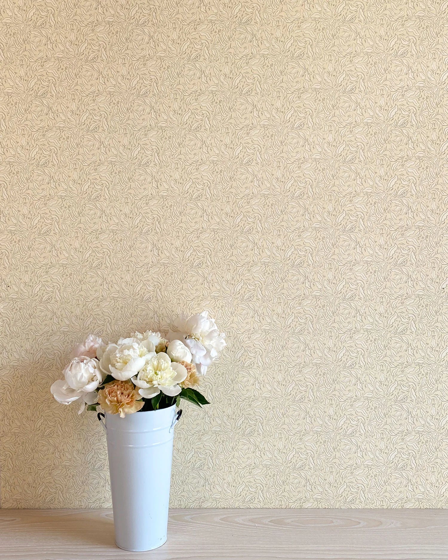 A vase of flowers stands in front of a wall papered in a painterly leaf print in tan and white on a cream field.